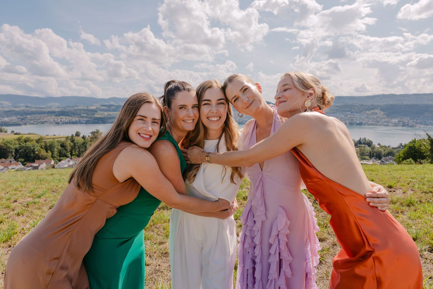 Five women in colorful dresses hugging outdoors with scenic lake and cloudy sky in background.