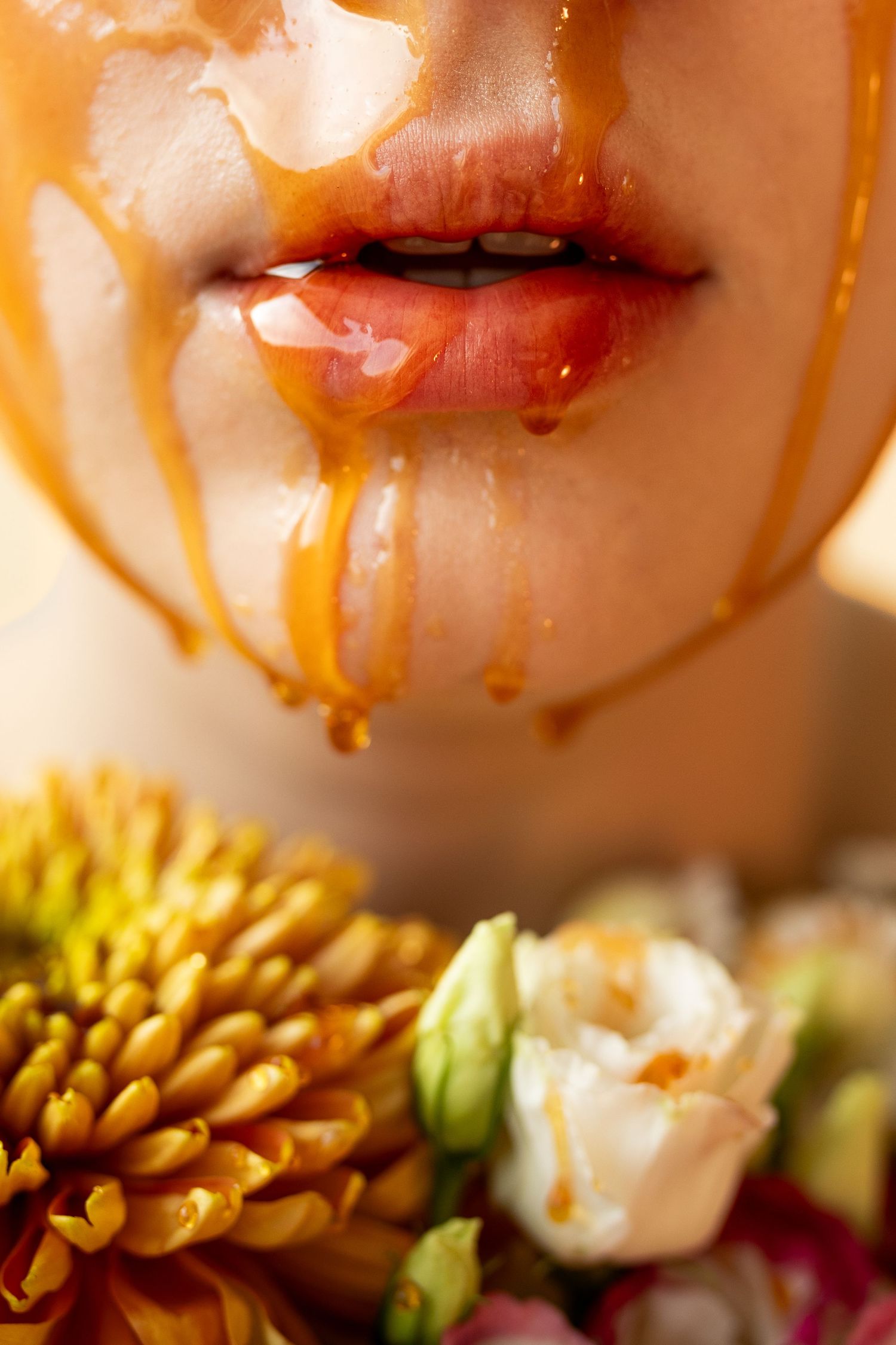 Close-up of a woman's face with golden honey dripping down her skin, surrounded by yellow chrysanthemums and white roses.