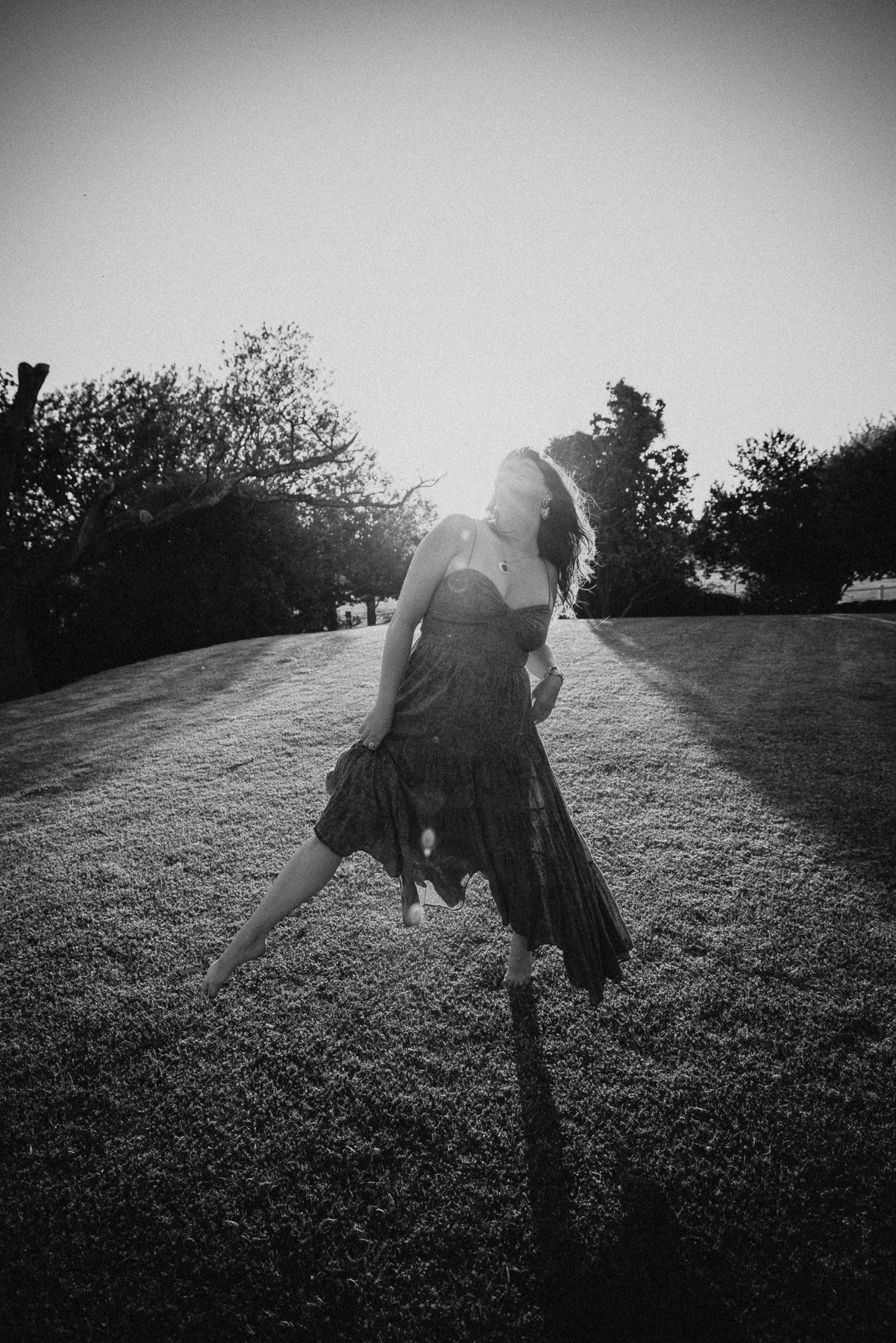 Black and white photo of a woman in a flowing dress dancing on a sunlit grass lawn with trees in the background.