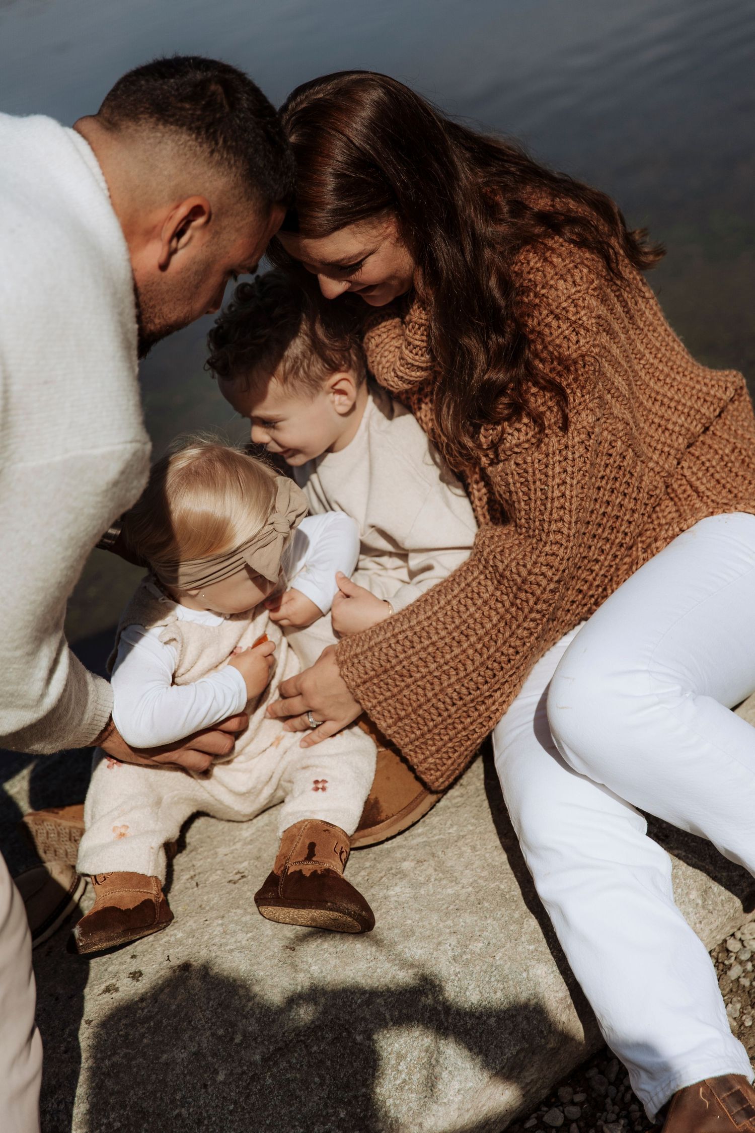 Family of four sitting together on rocks by a lake, parents bonding with their two young children.