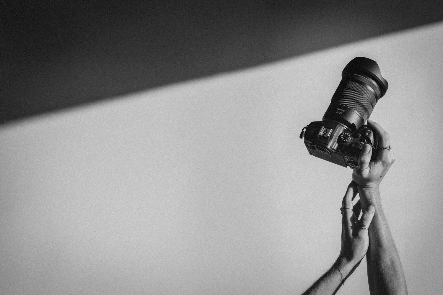 Black and white photo of a hand holding a DSLR camera with a large lens raised against a plain wall.
