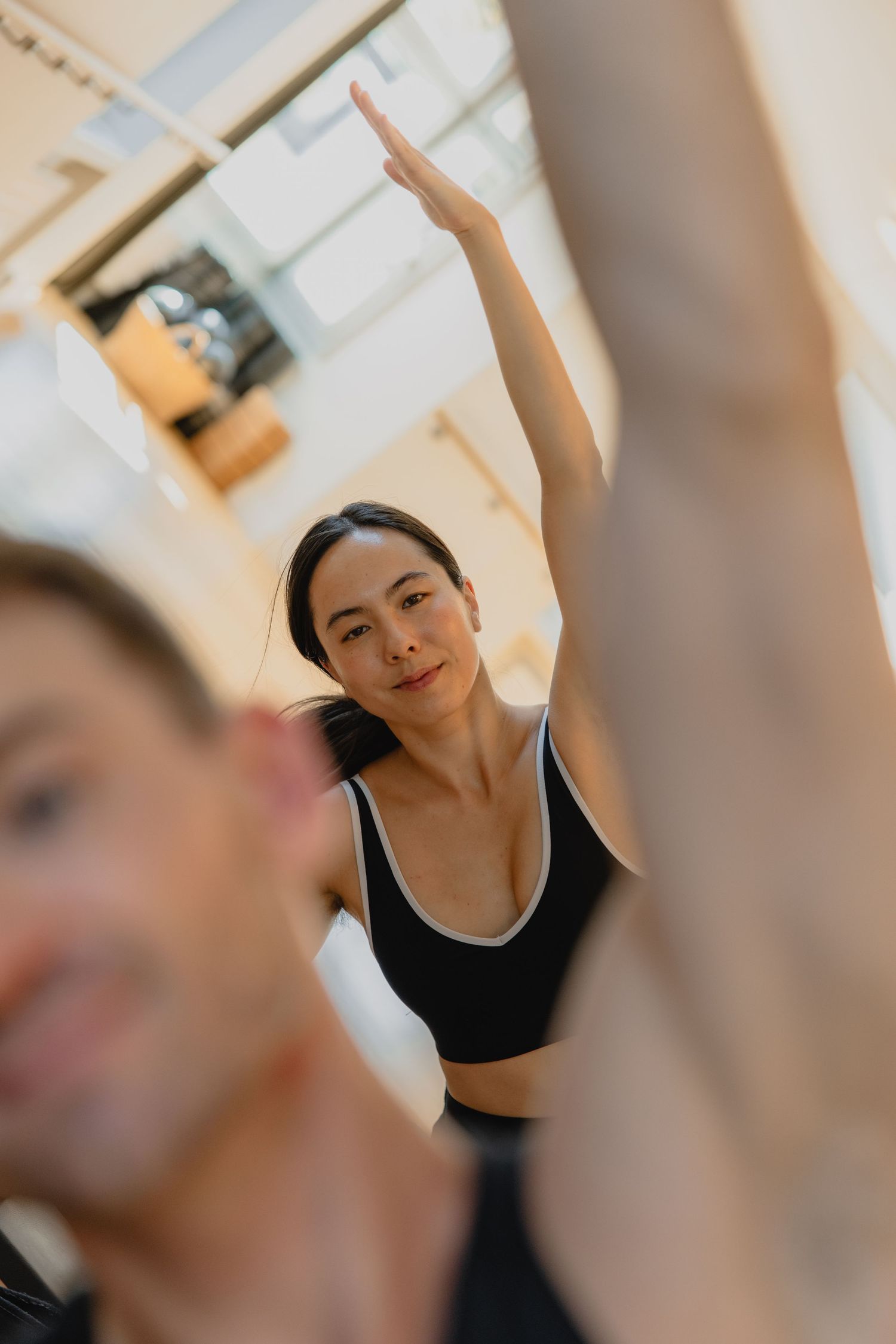Woman in black sports bra raises arm during yoga class with instructor in bright studio.