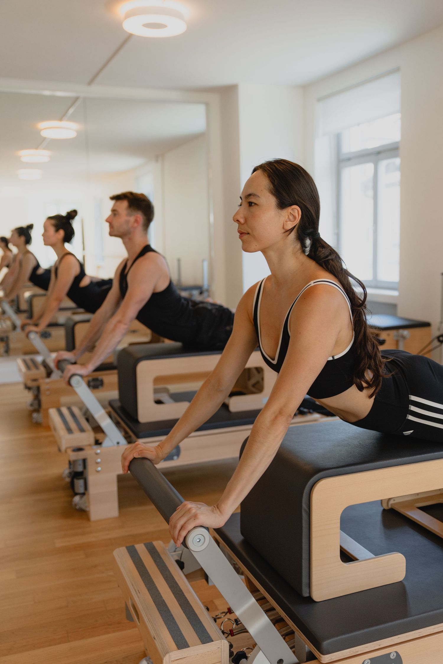 Group of people doing Pilates reformer exercises in a bright modern studio with wooden equipment.