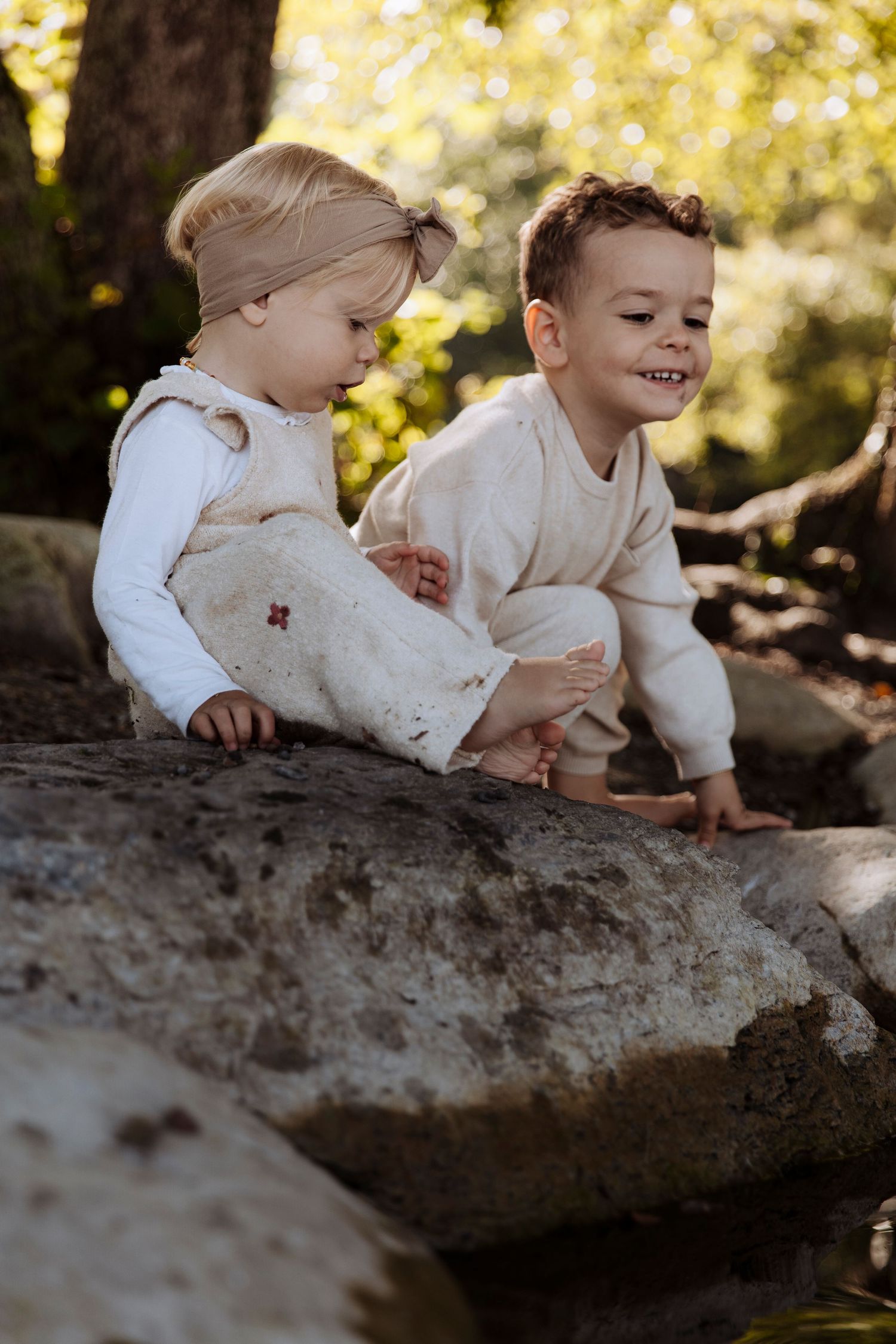 Two young siblings in neutral outfits playing together on rocks in a sunlit forest setting.