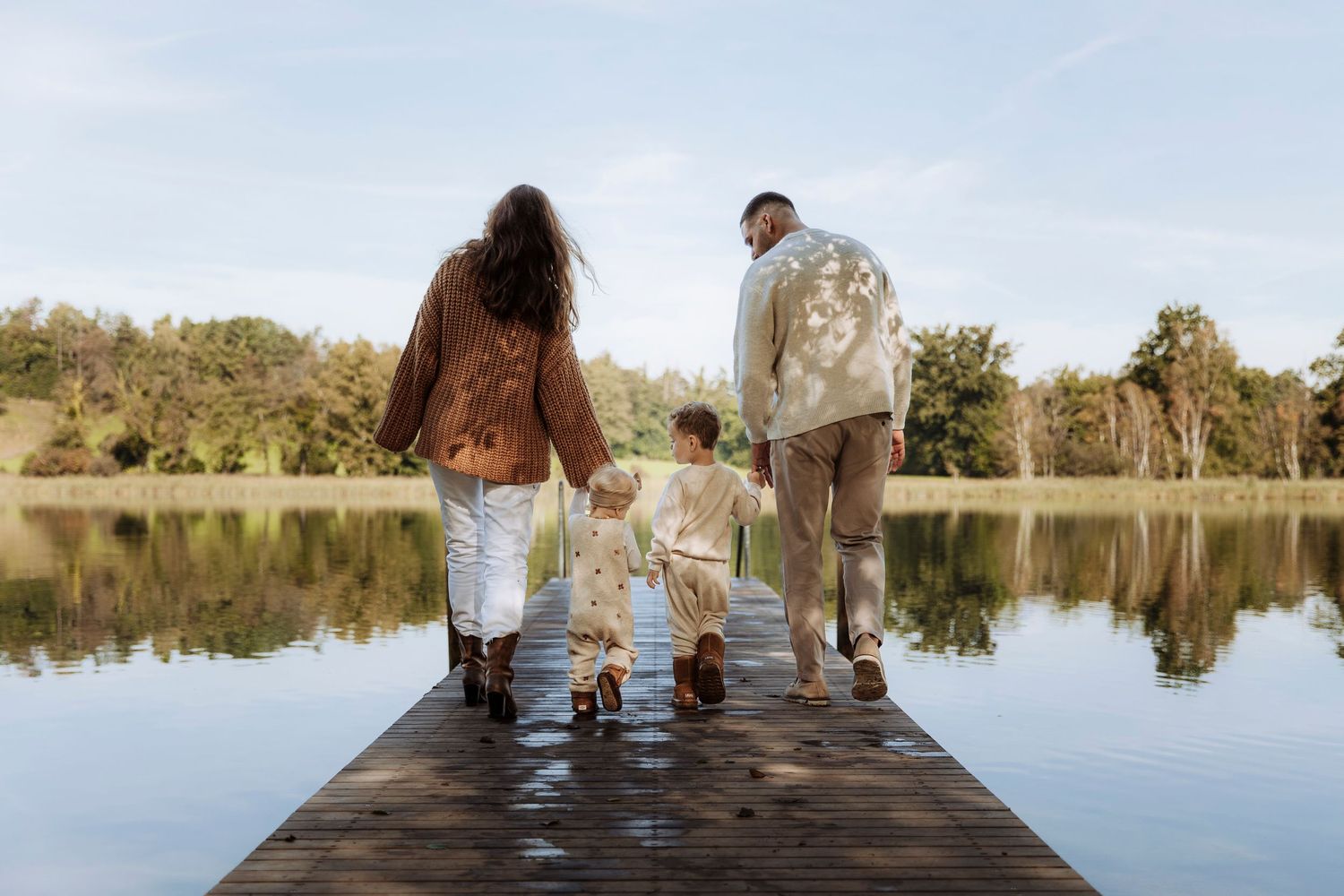 Family of three walking hand-in-hand on a wooden dock toward a peaceful reflective lake.