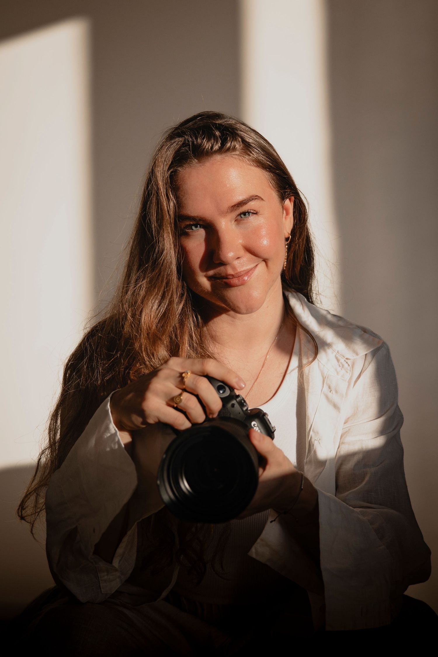 Young woman in white shirt smiling warmly while holding a DSLR camera in soft natural window light.