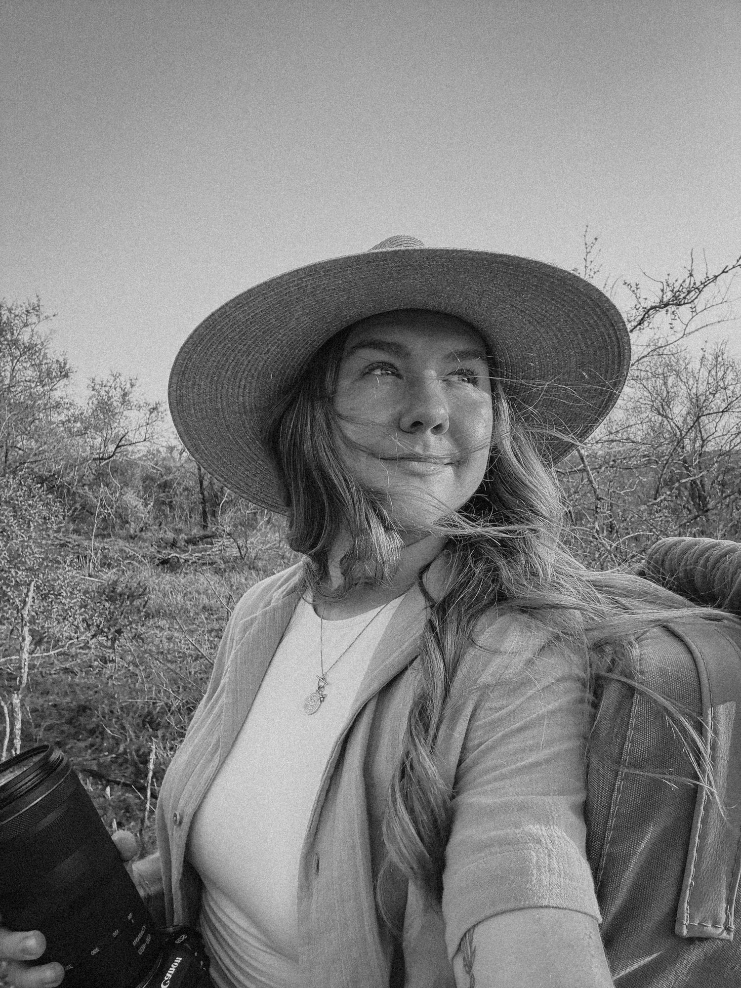 Woman in wide-brim hat holding camera lens outdoors in arid landscape, black and white portrait photo.
