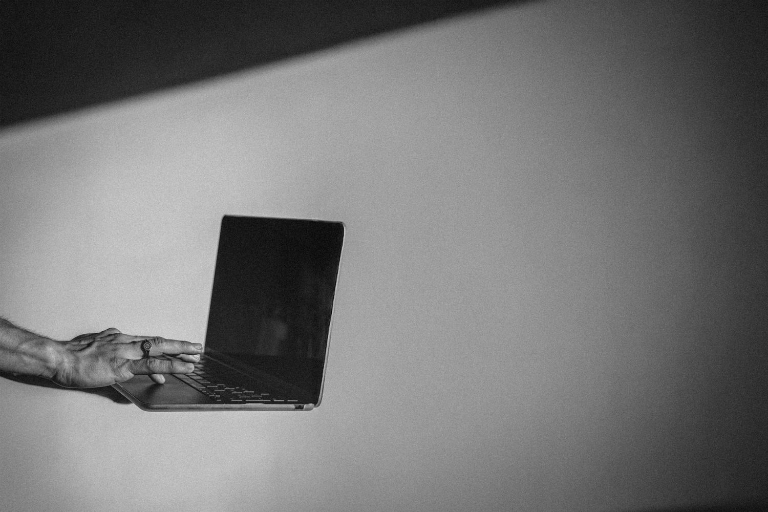A hand holds an open laptop against a plain light background in a dramatic black and white photo.