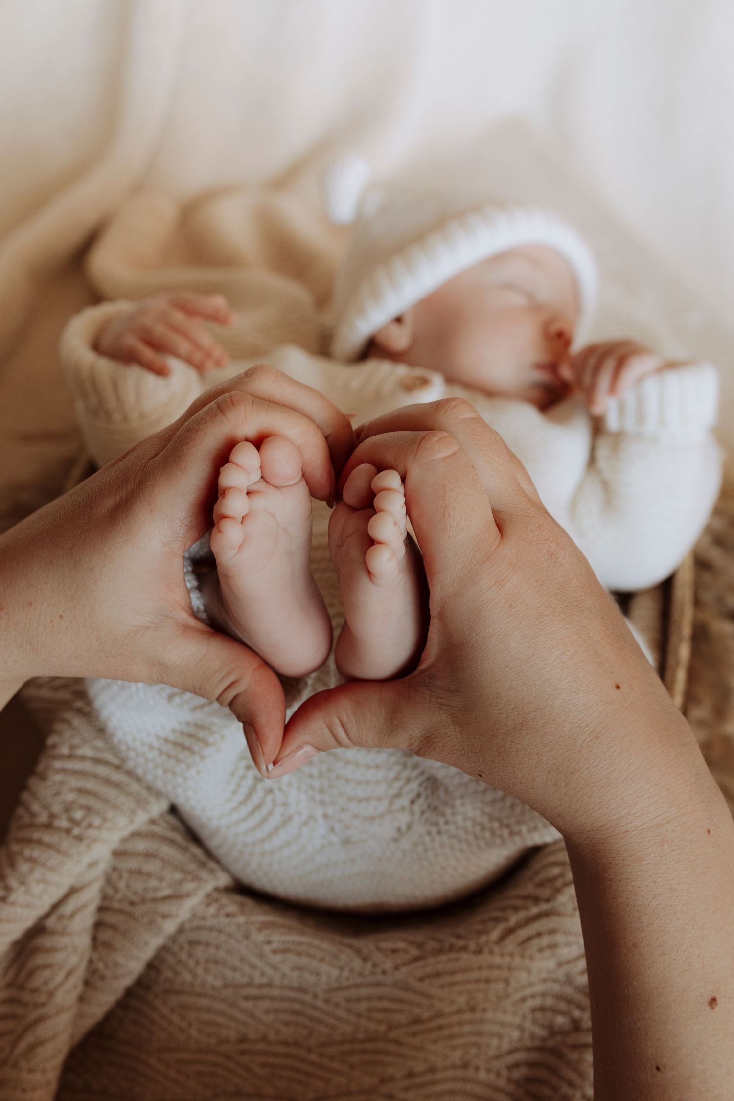 Adult hands forming a heart shape around tiny newborn baby feet while infant sleeps on a knit blanket.