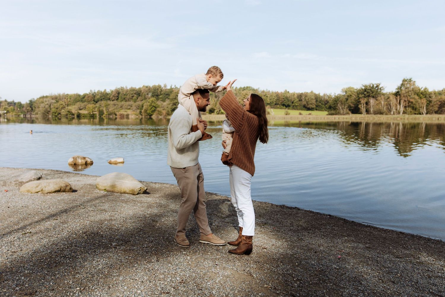Family of three enjoying a sunny day by a calm lake, with parents lifting their baby joyfully.