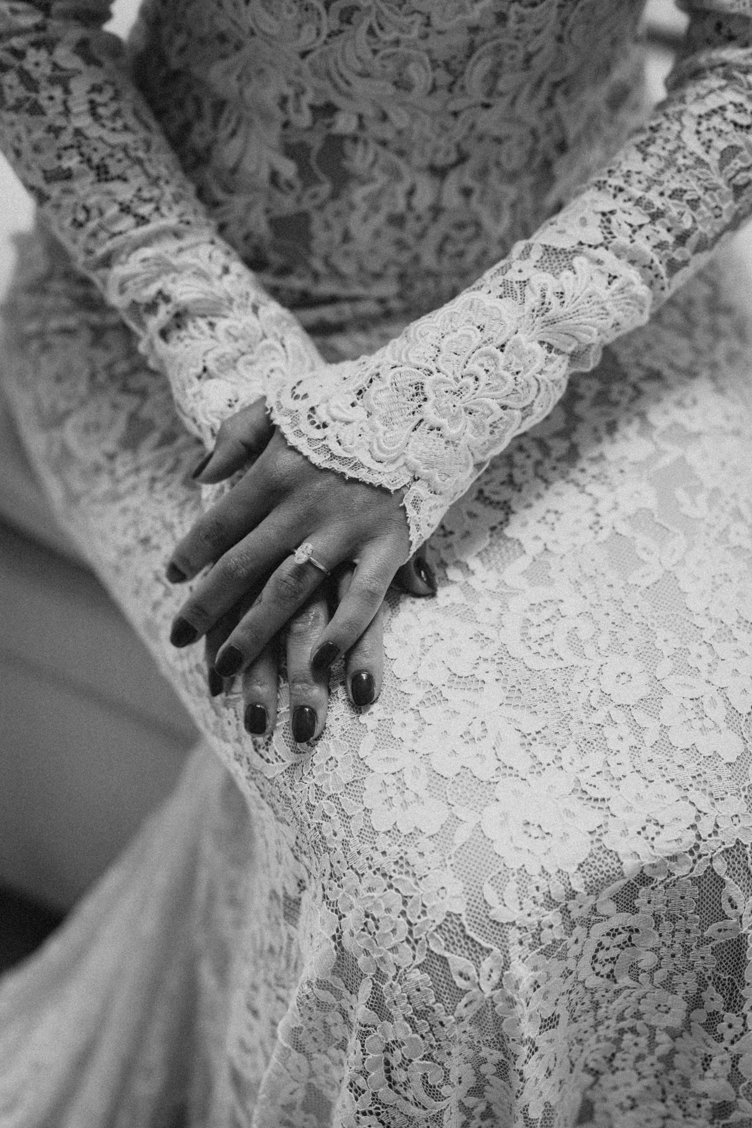 Close-up black and white photo of bride's hands with dark nail polish and ring on lace wedding dress.