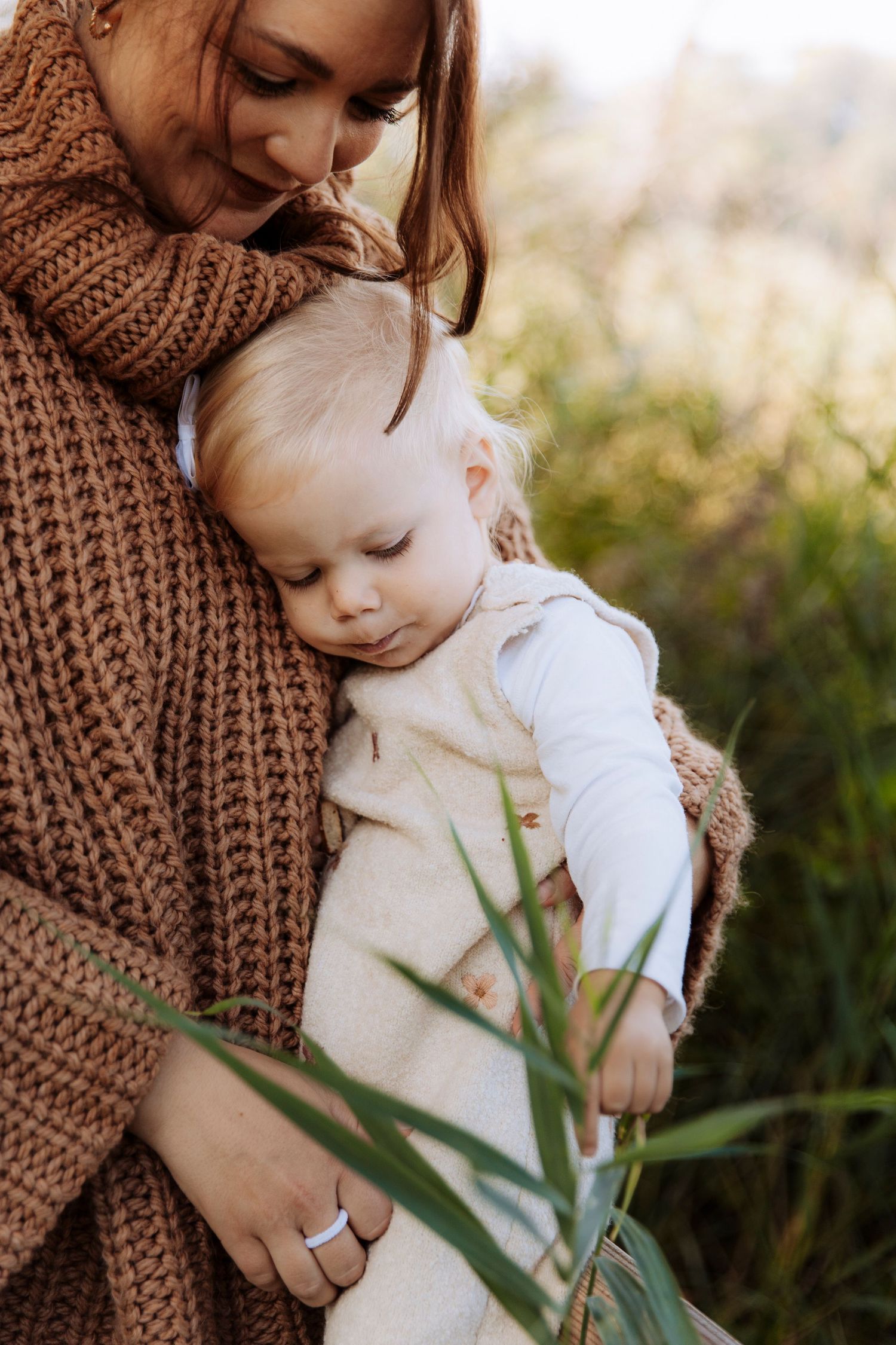 A mother in a brown knit cardigan tenderly holds her sleeping blonde baby outdoors in a green field.