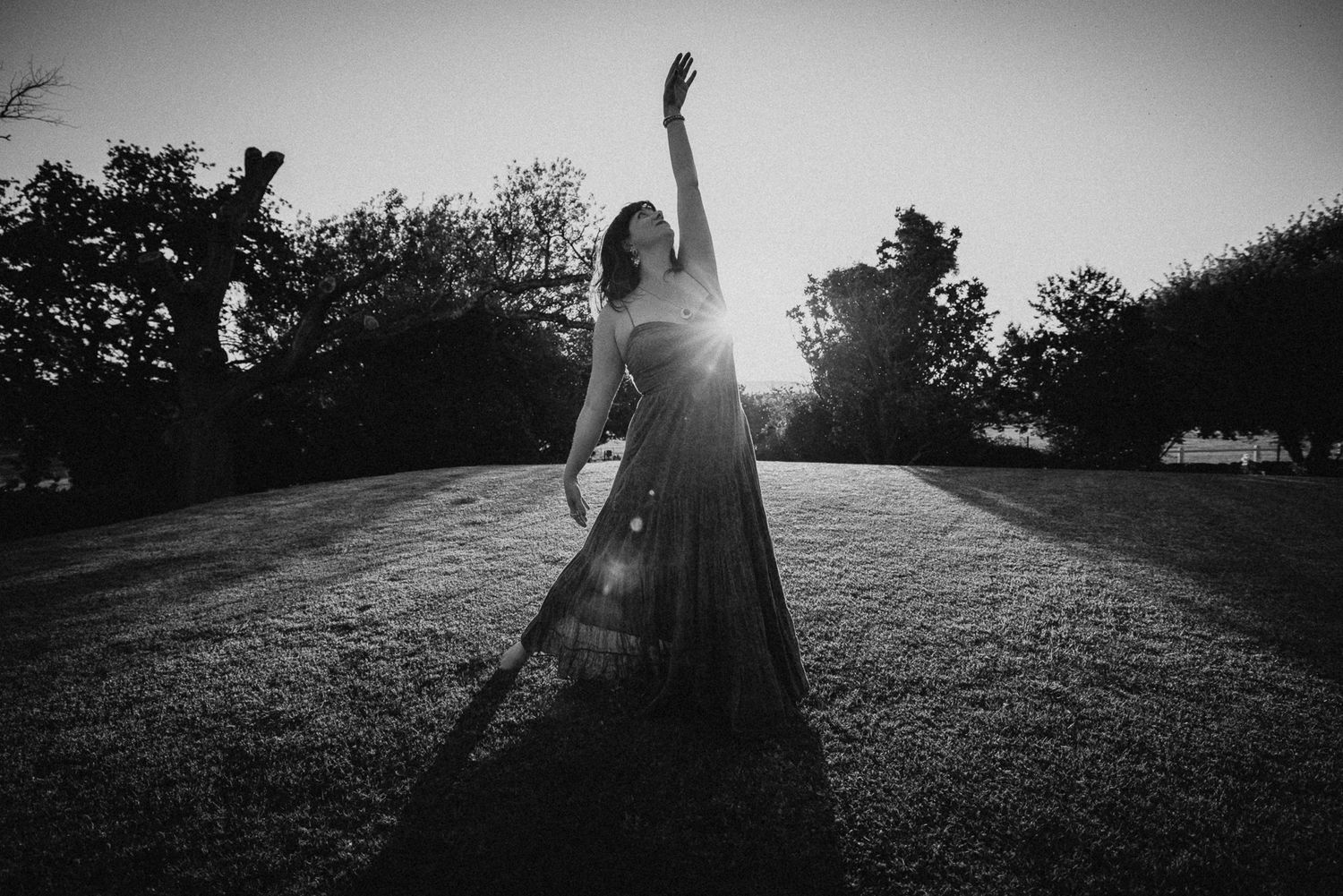 Black and white photo of a woman in a flowing gown dancing outdoors at sunset with one arm raised.