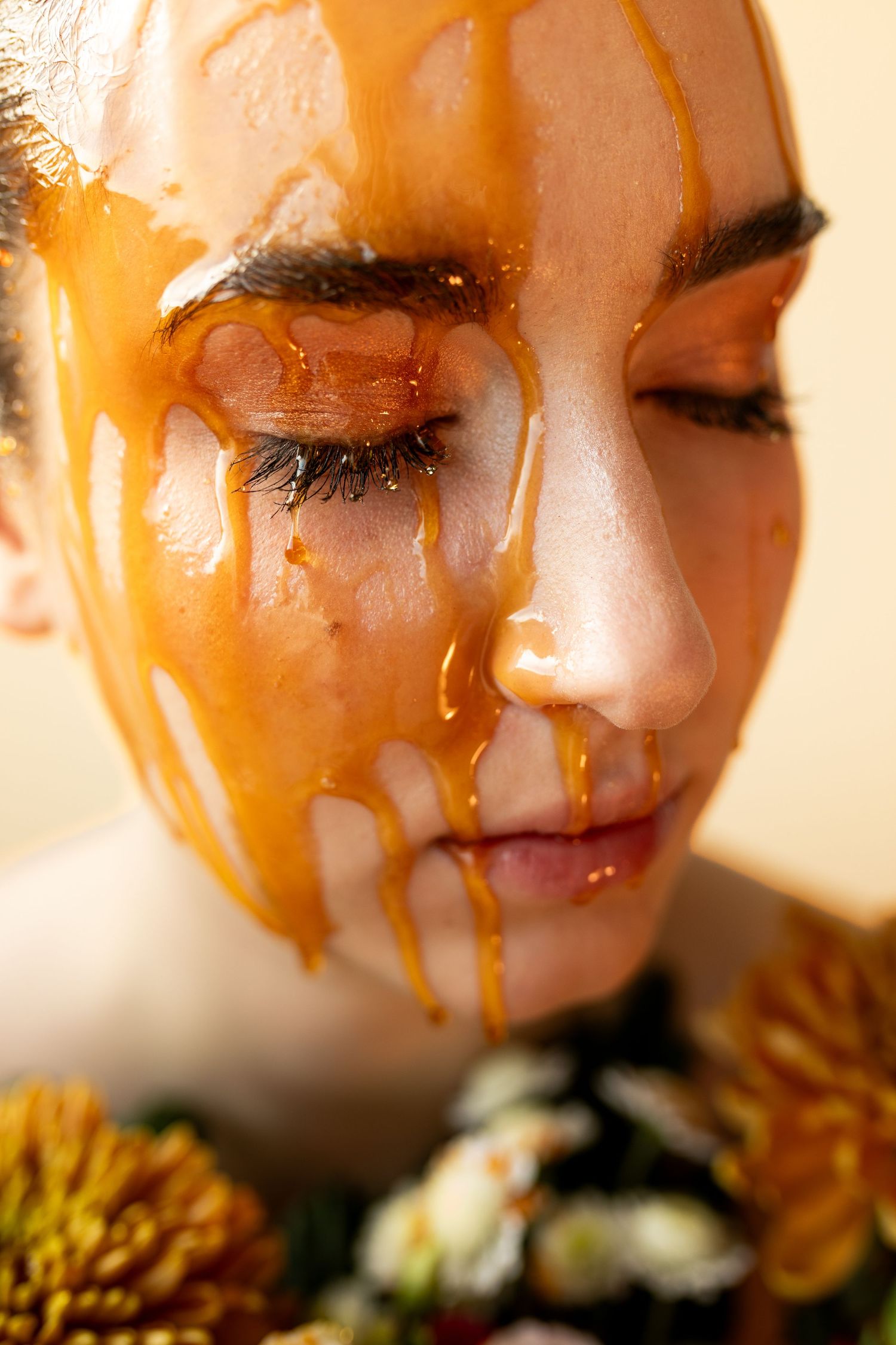 Close-up of a woman with golden honey dripping down her face, eyes closed, with yellow flowers at her chest.