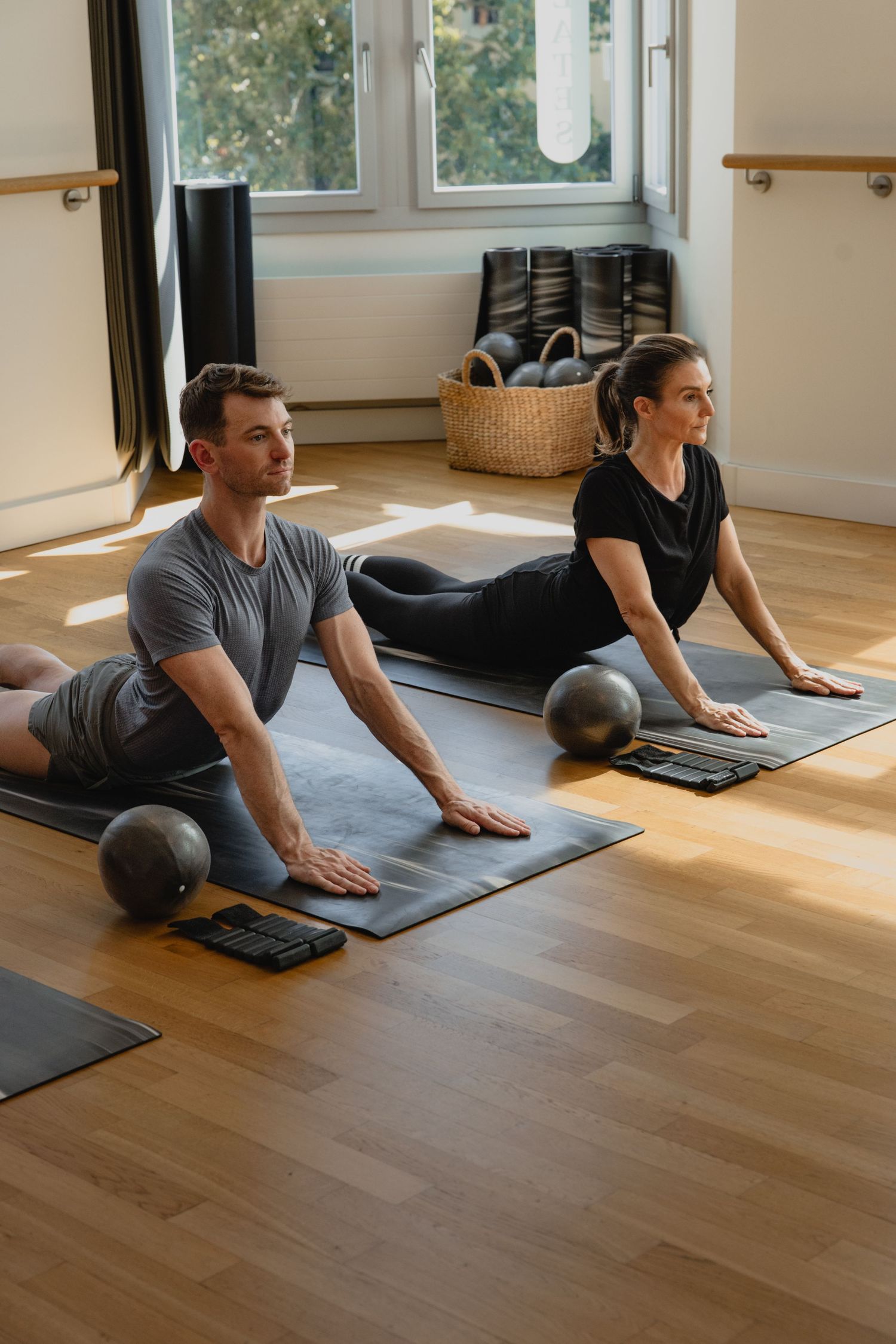 A man and woman practice pilates exercises on gray mats in a sunlit studio with wooden floors and balance balls nearby.
