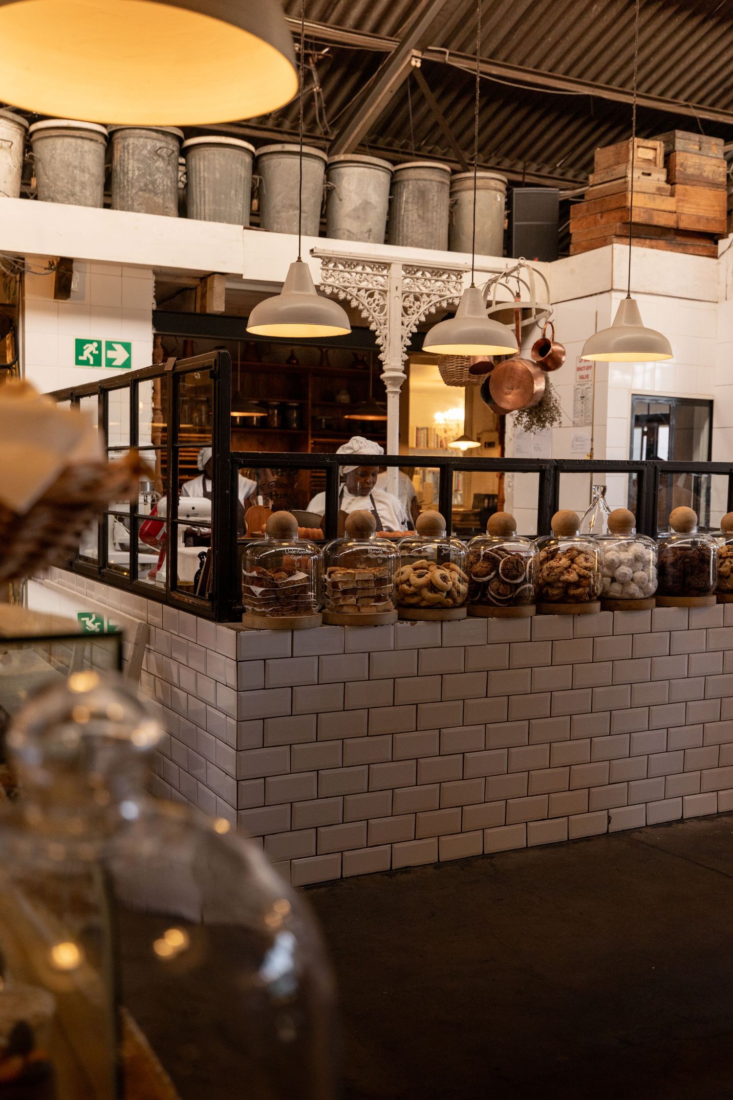 Rustic bakery counter with glass jars filled with cookies and pastries, pendant lights, white subway tiles, and vintage metal bins.