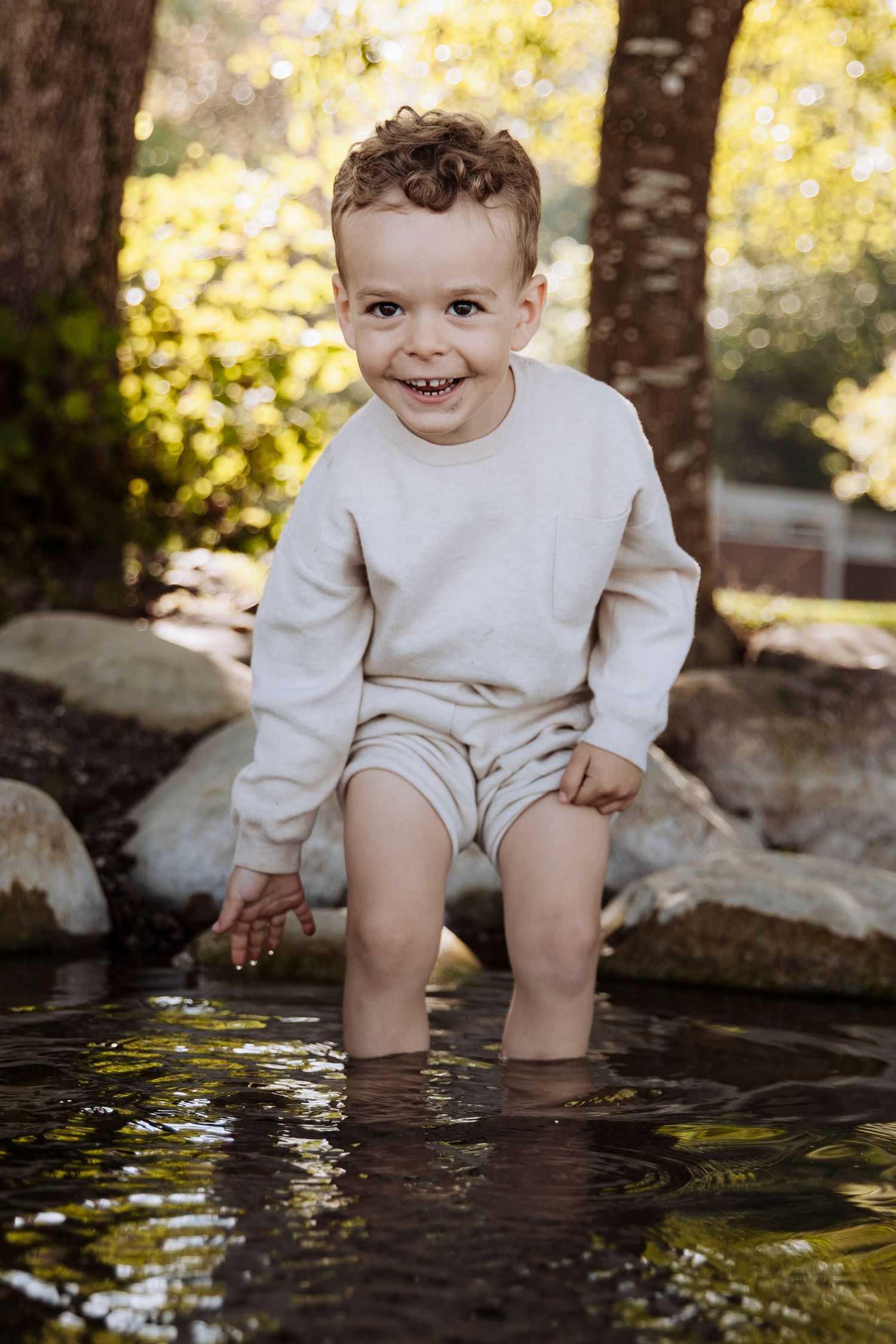 Happy toddler boy in beige outfit standing in shallow creek water outdoors.
