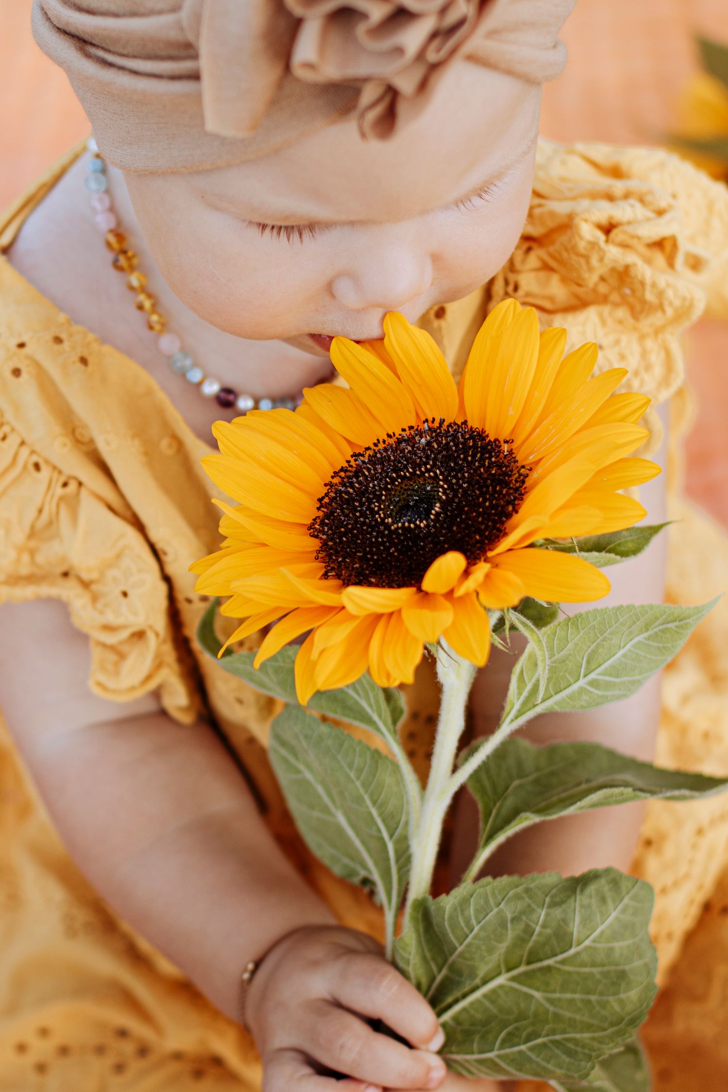 Sweet baby girl in yellow dress and tan turban smelling a large bright yellow sunflower she is holding.