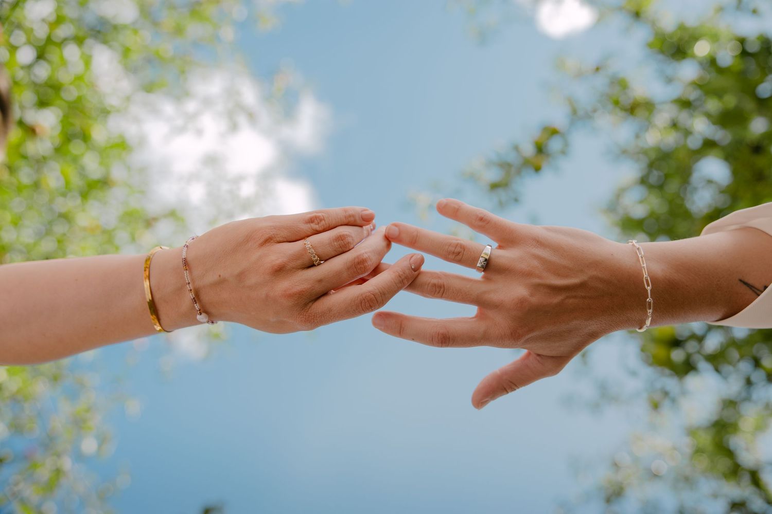 Two hands reaching toward each other wearing gold rings and bracelets against a blue sky with green trees.