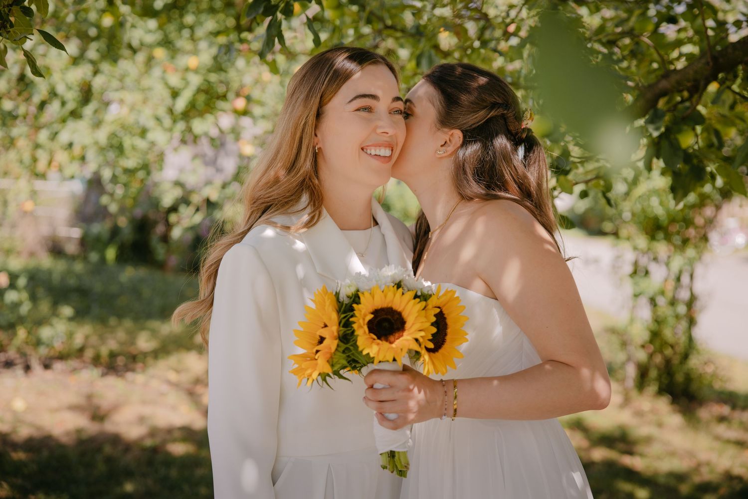 Two brides in white dresses share a tender kiss while holding a vibrant sunflower bouquet outdoors.