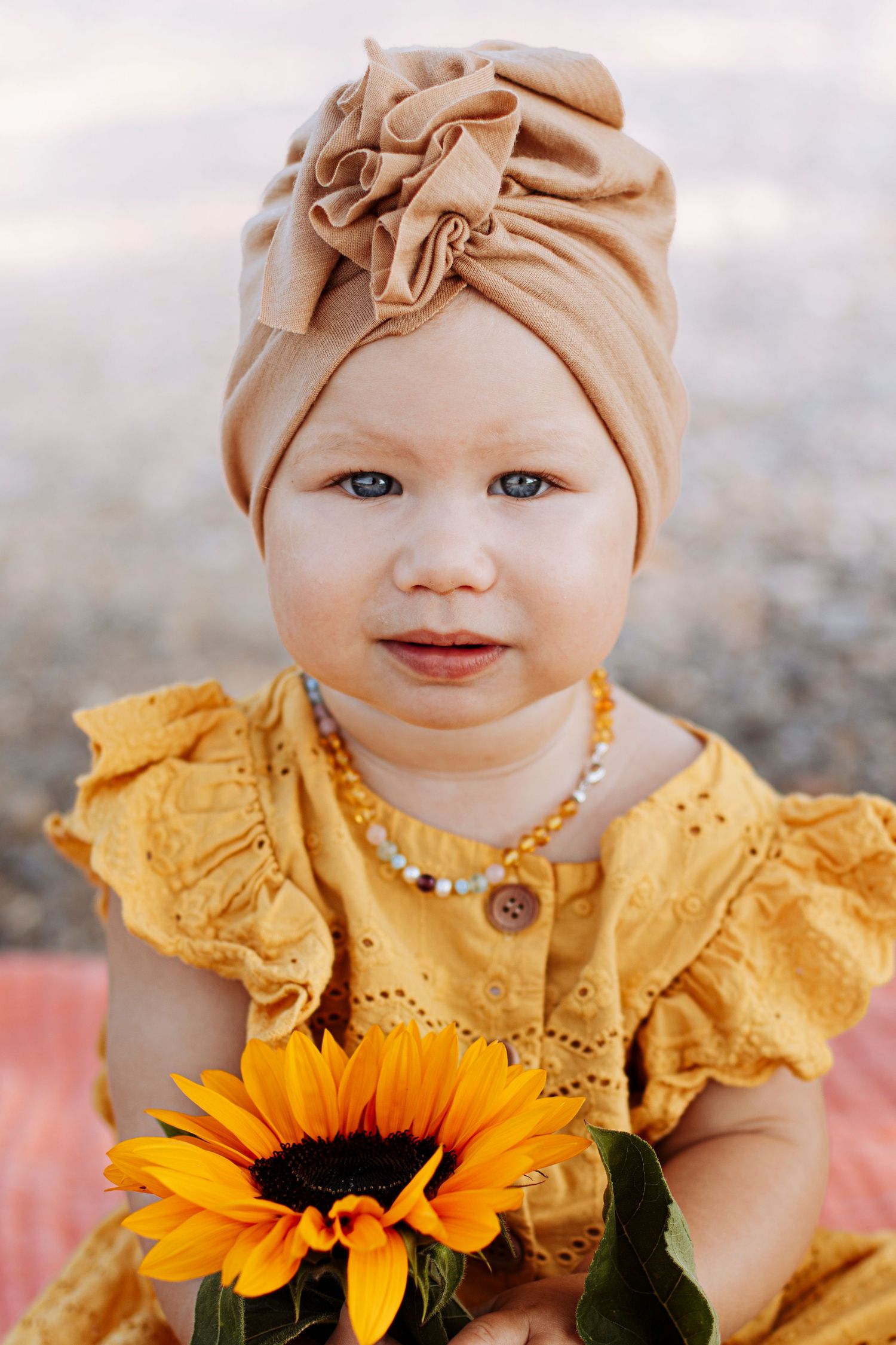 Adorable baby girl in yellow dress and tan turban headband holding a bright sunflower outdoors.