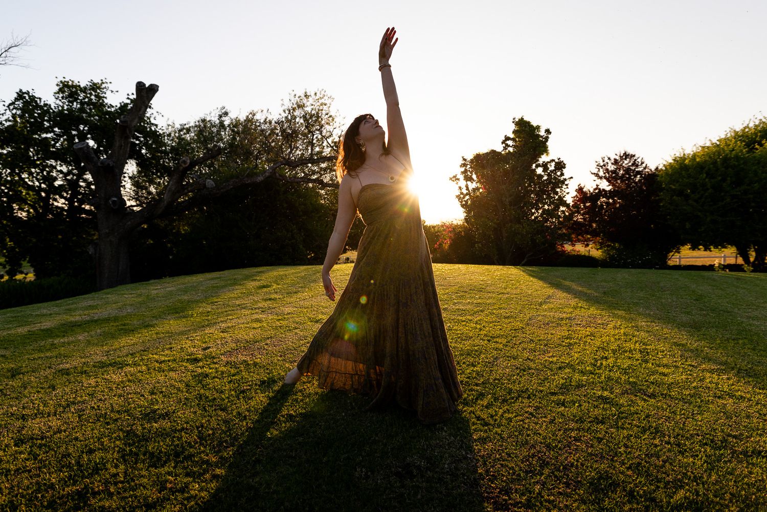 Woman in flowing dress dancing with raised arm in golden sunset light on green lawn.