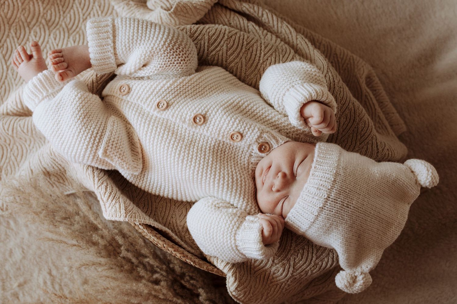 A newborn baby in a cream knit suit and hat sleeps peacefully in a wicker basket.