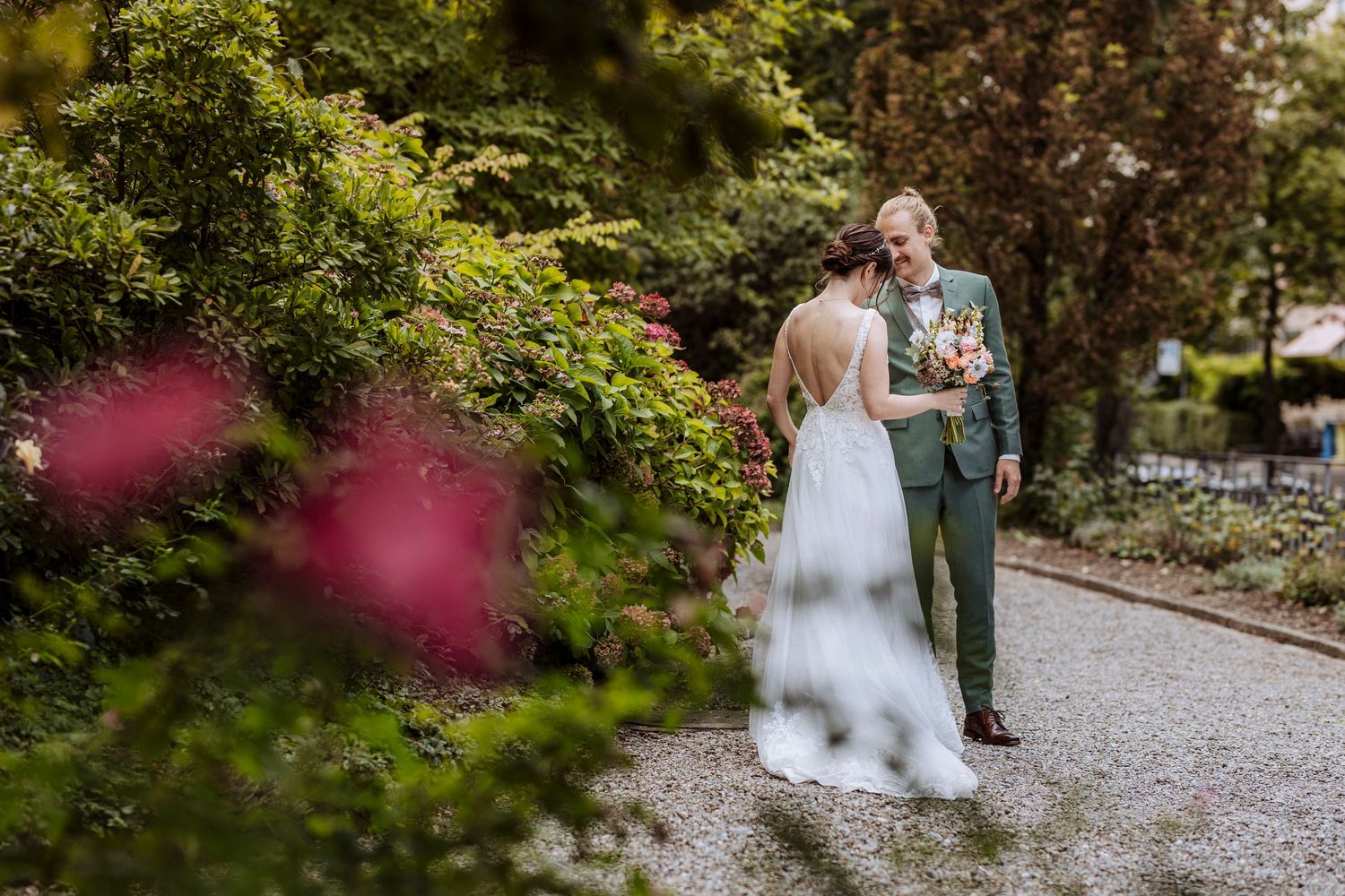 Bride and groom share a romantic moment on a gravel path surrounded by lush green and pink garden foliage.