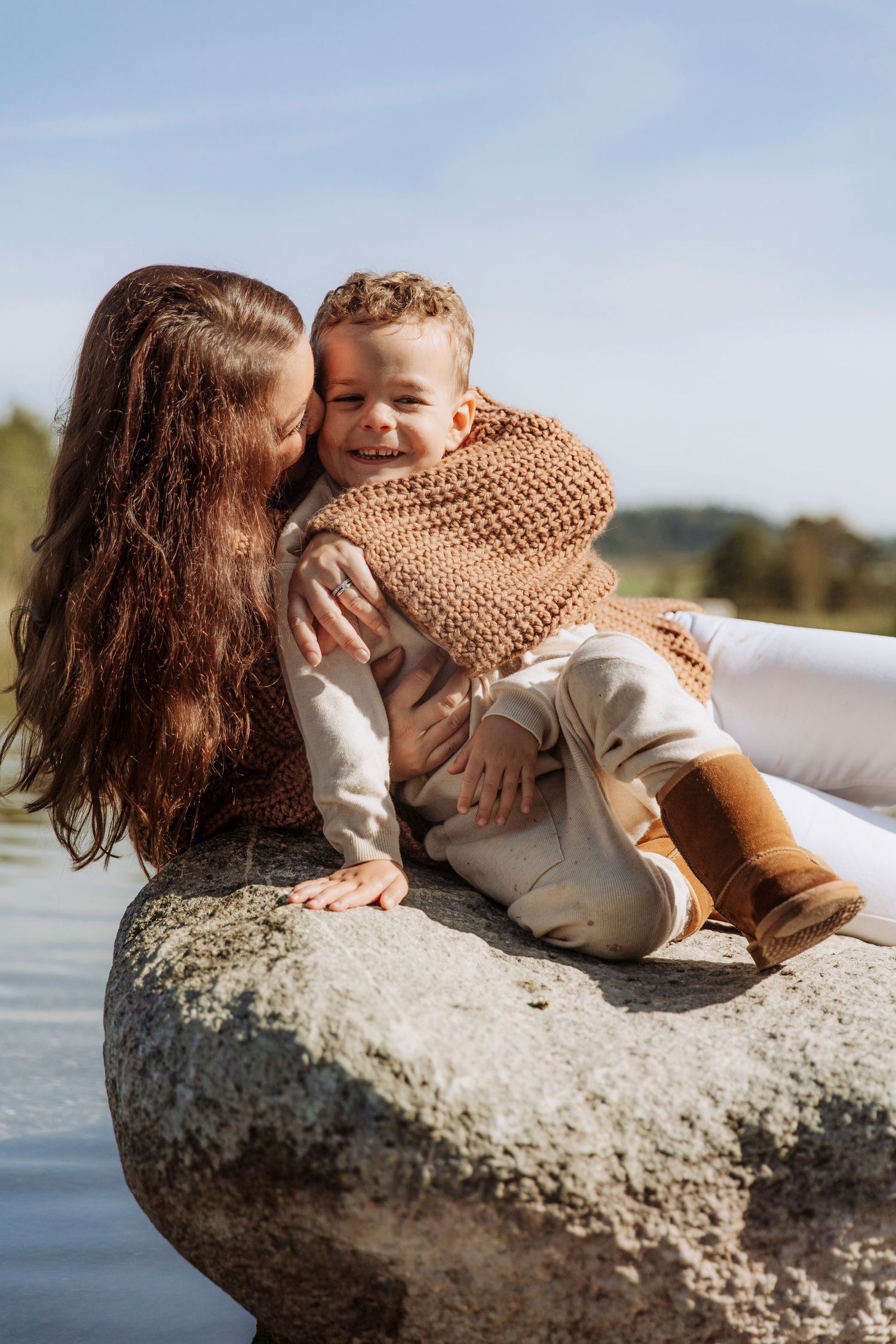 Mother in brown knit sweater cuddling her laughing toddler son on a large rock by a lake.