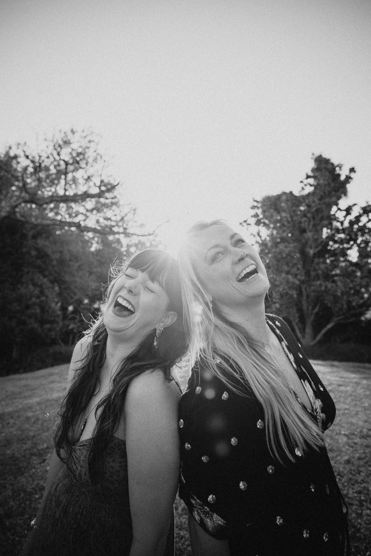 Two women laughing joyfully back-to-back outdoors in a sunlit black and white portrait photo.