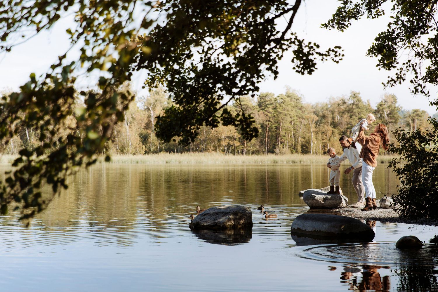 A family enjoys a sunny day standing on rocks in a calm lake surrounded by trees.