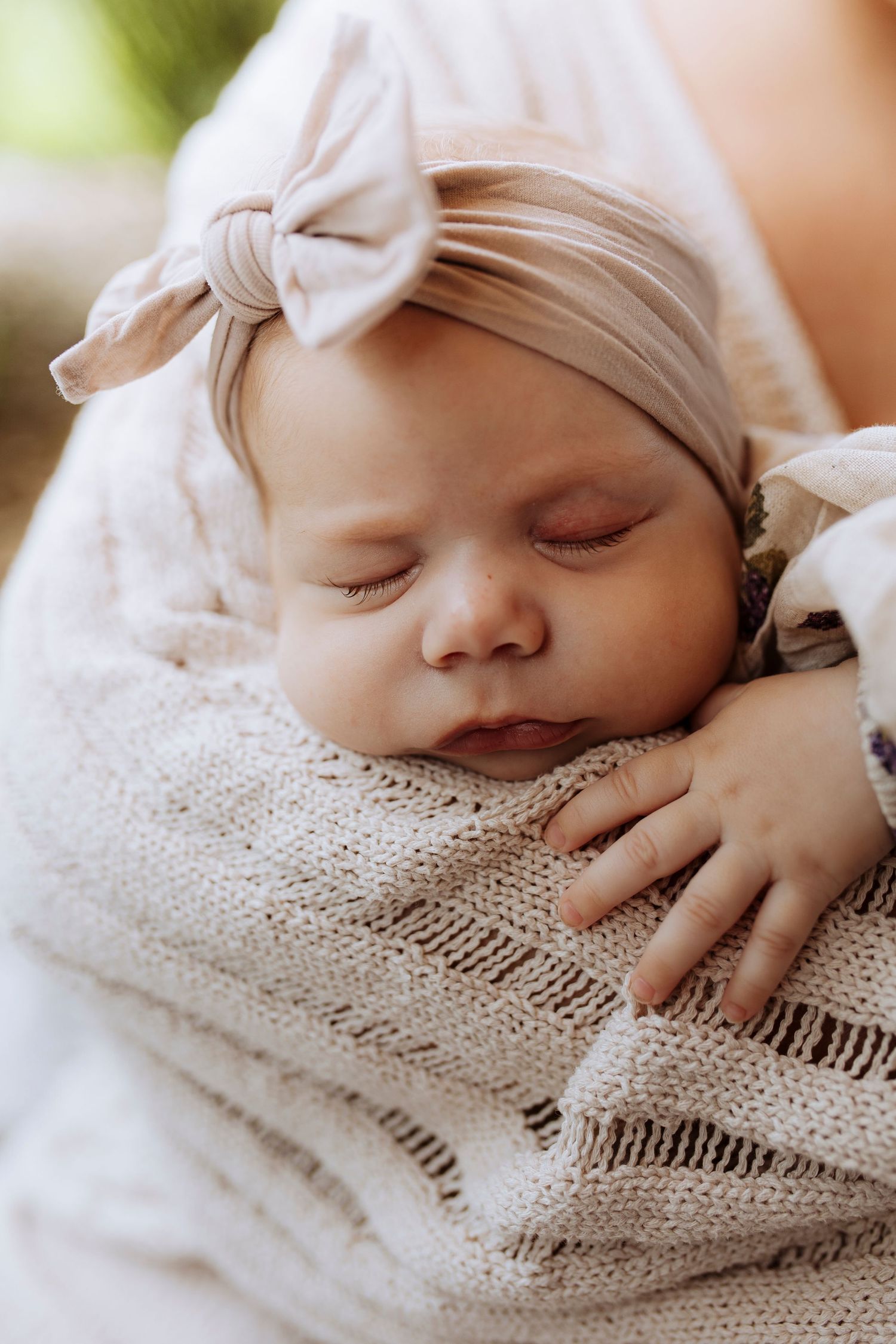 A sleeping newborn baby girl with a bow headband is cradled in soft knit blanket.