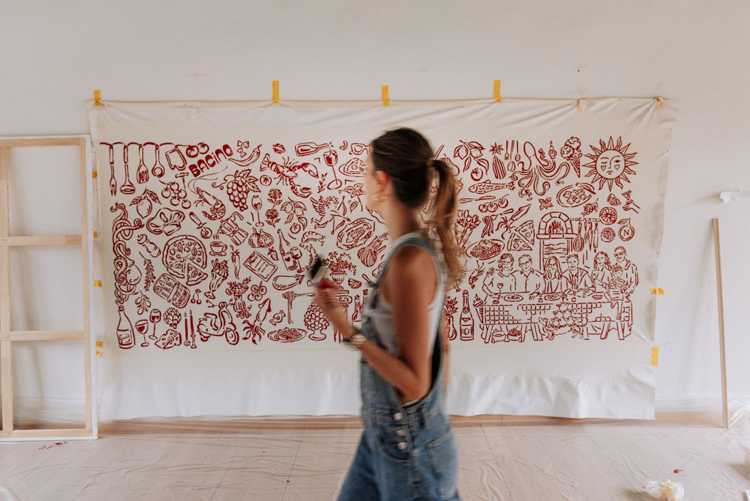 Artist in denim overalls standing before a large white canvas with red doodle artwork on wall.