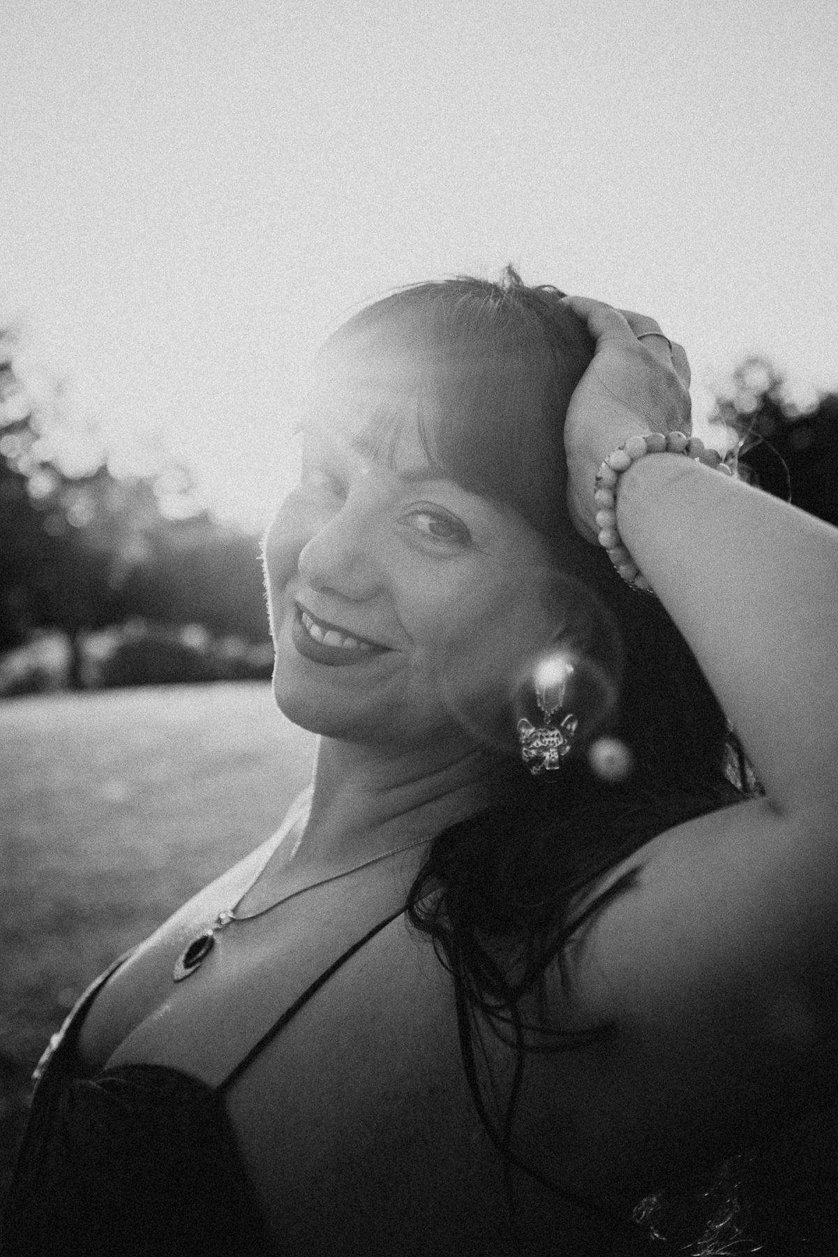 Black and white portrait of a smiling woman with hand in hair, wearing earrings and necklace, backlit by sunset.