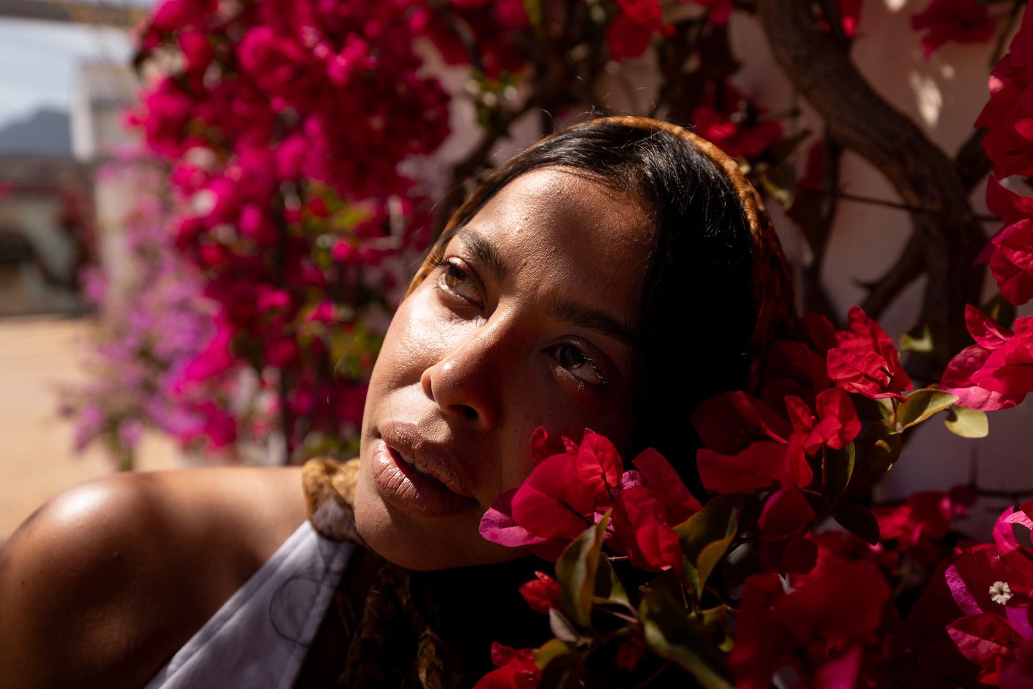Young woman with closed eyes resting among vibrant red bougainvillea flowers in bright sunlight.