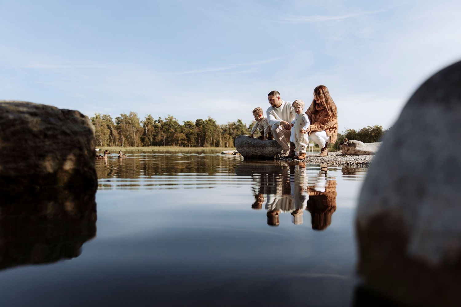 Family of three standing on rocks by a tranquil lake on a bright sunny day.