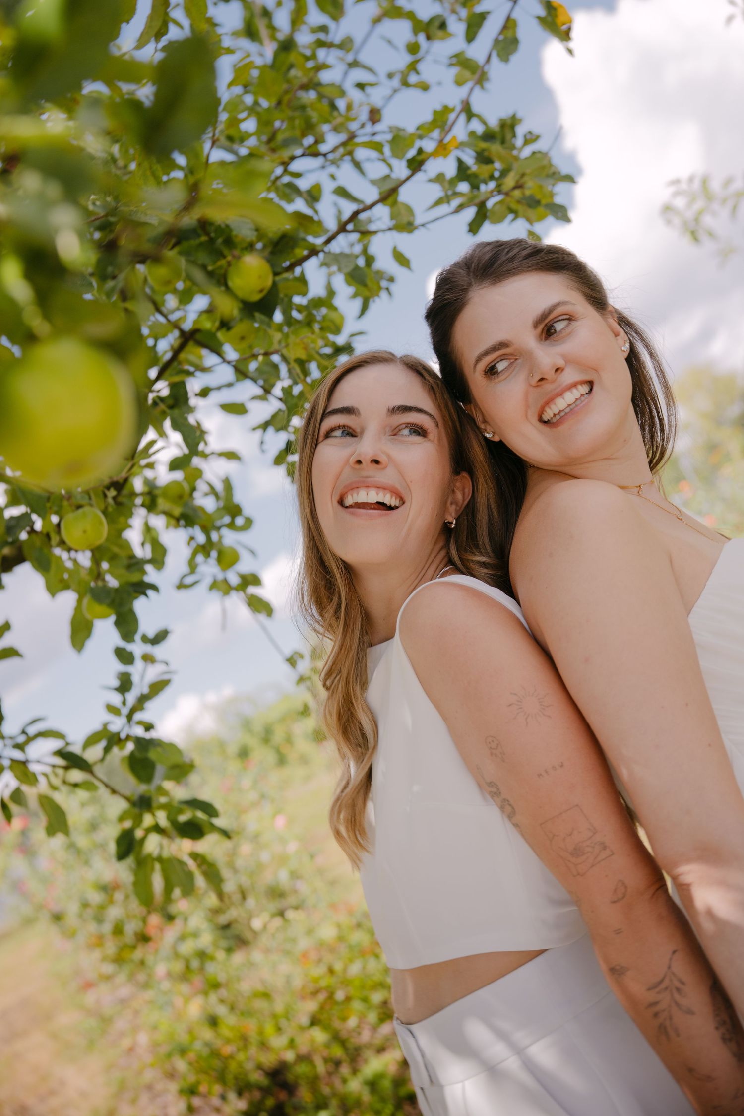 Two smiling women in white dresses stand together outdoors near a green apple tree on a sunny day.