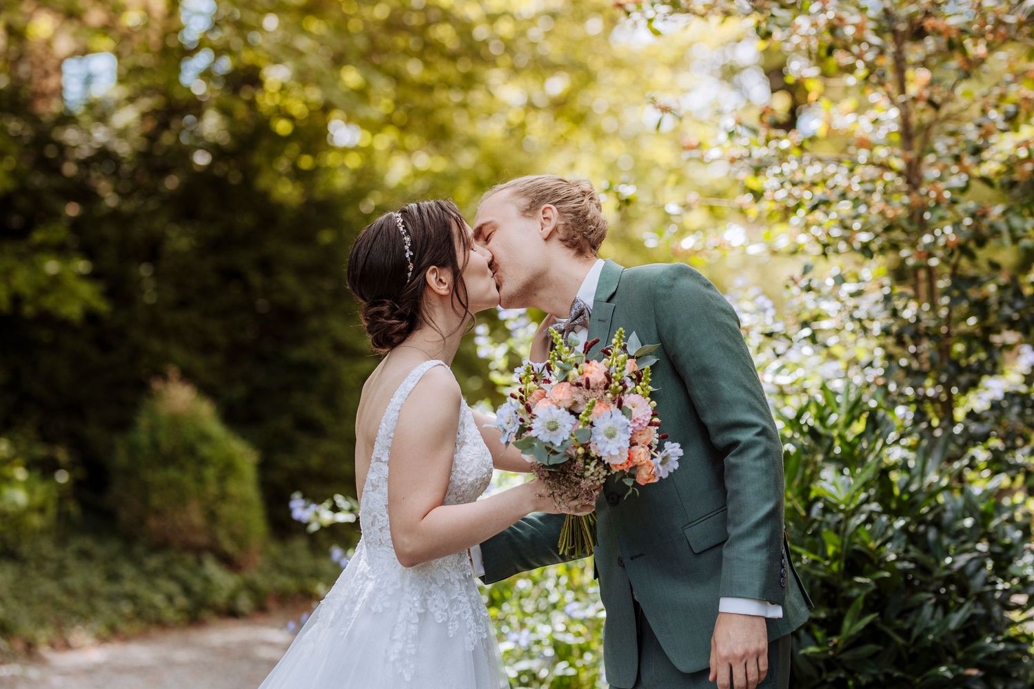 A newlywed couple shares a romantic kiss while the groom holds a pastel bouquet in a lush garden.