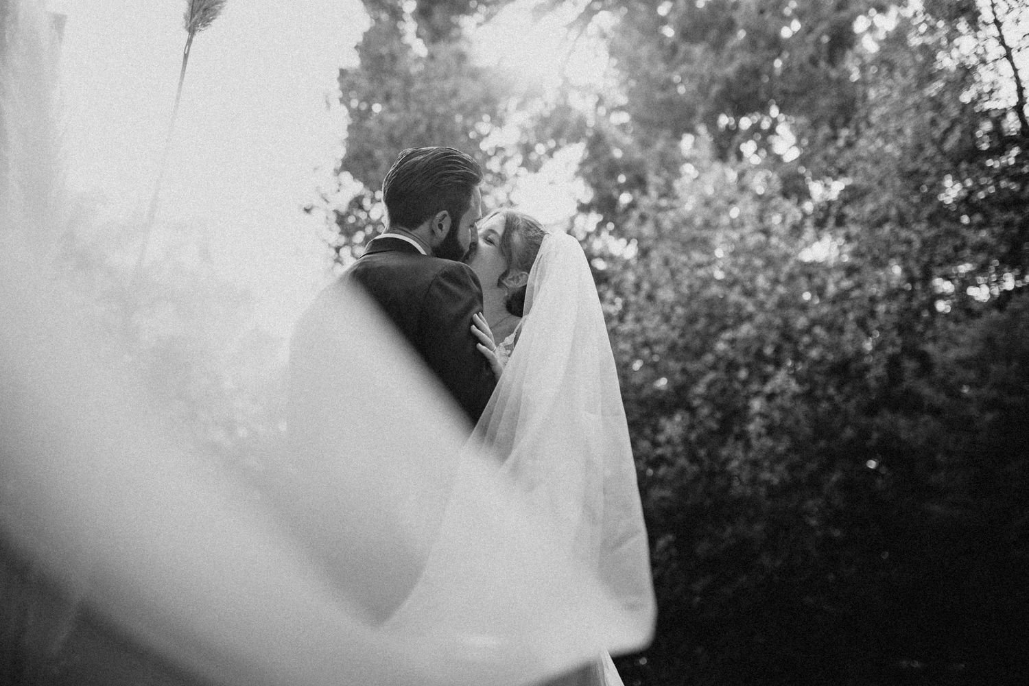 A bride and groom embrace outdoors with a flowing veil in a romantic black and white wedding photo.