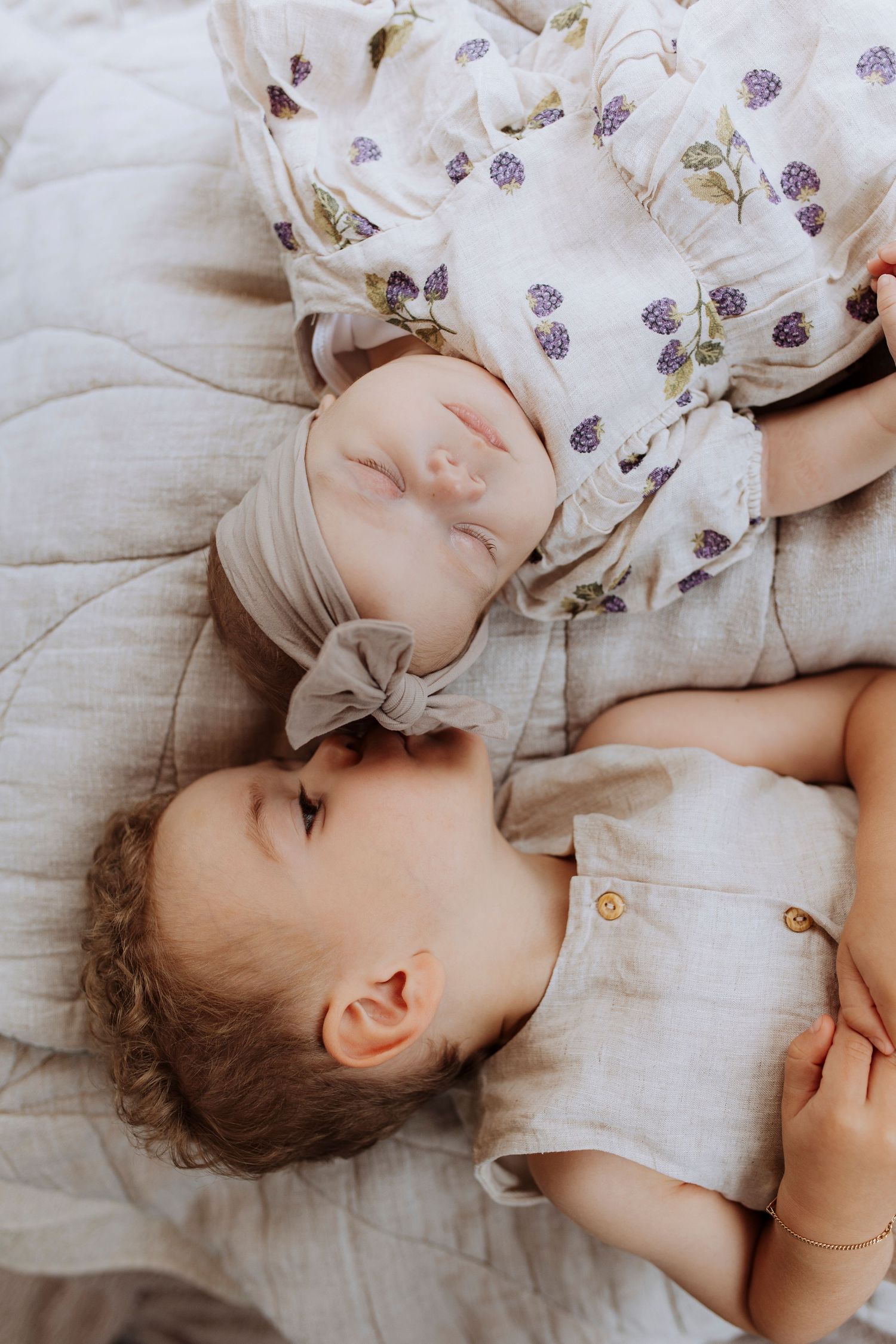 A toddler in a linen romper lies beside a sleeping newborn baby in a floral outfit.