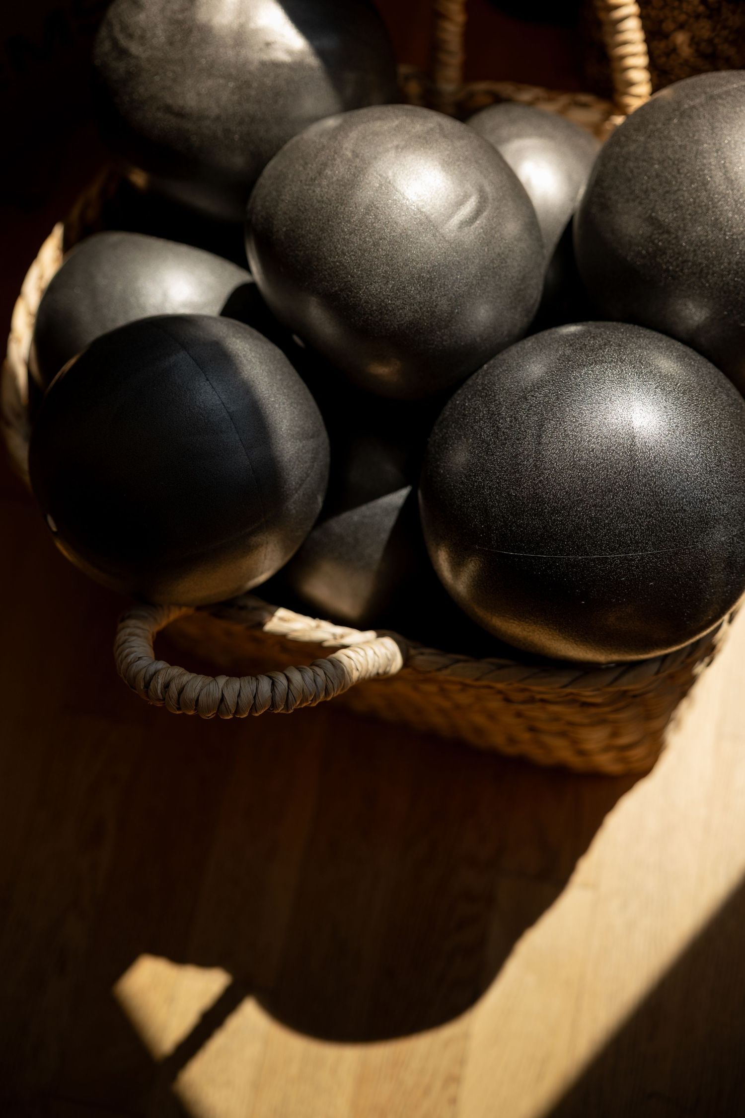 Dark metal bocce balls stored in a woven wicker basket on a wooden floor in warm sunlight.