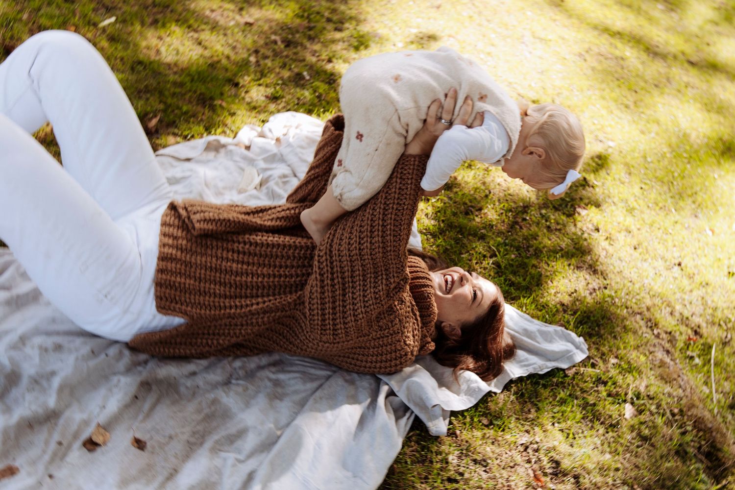 A joyful mother lies on a blanket on grass playfully lifting her baby into the air on a sunny day.