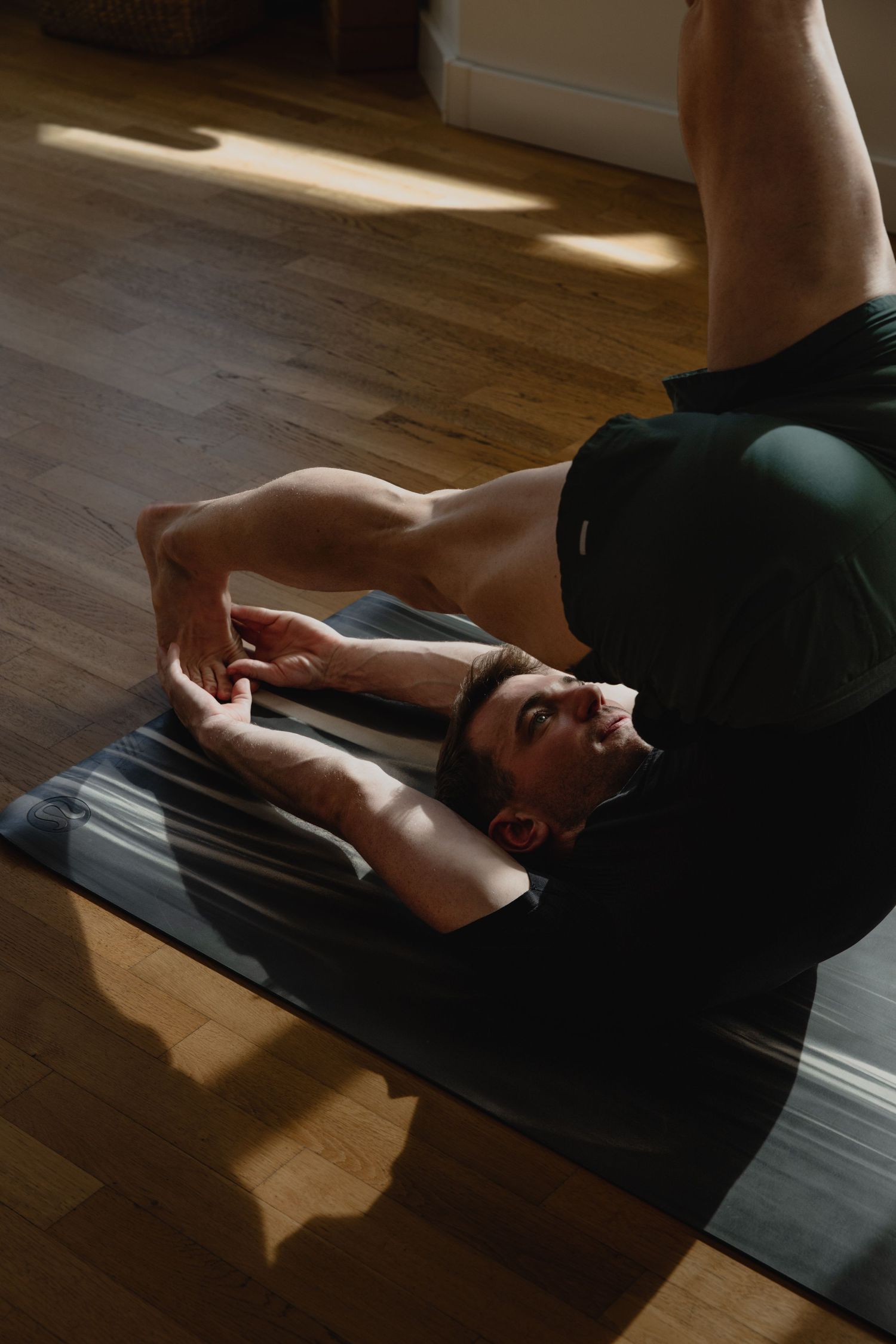 Man practicing yoga on a mat indoors, performing a leg raise exercise in natural sunlight on hardwood floor.
