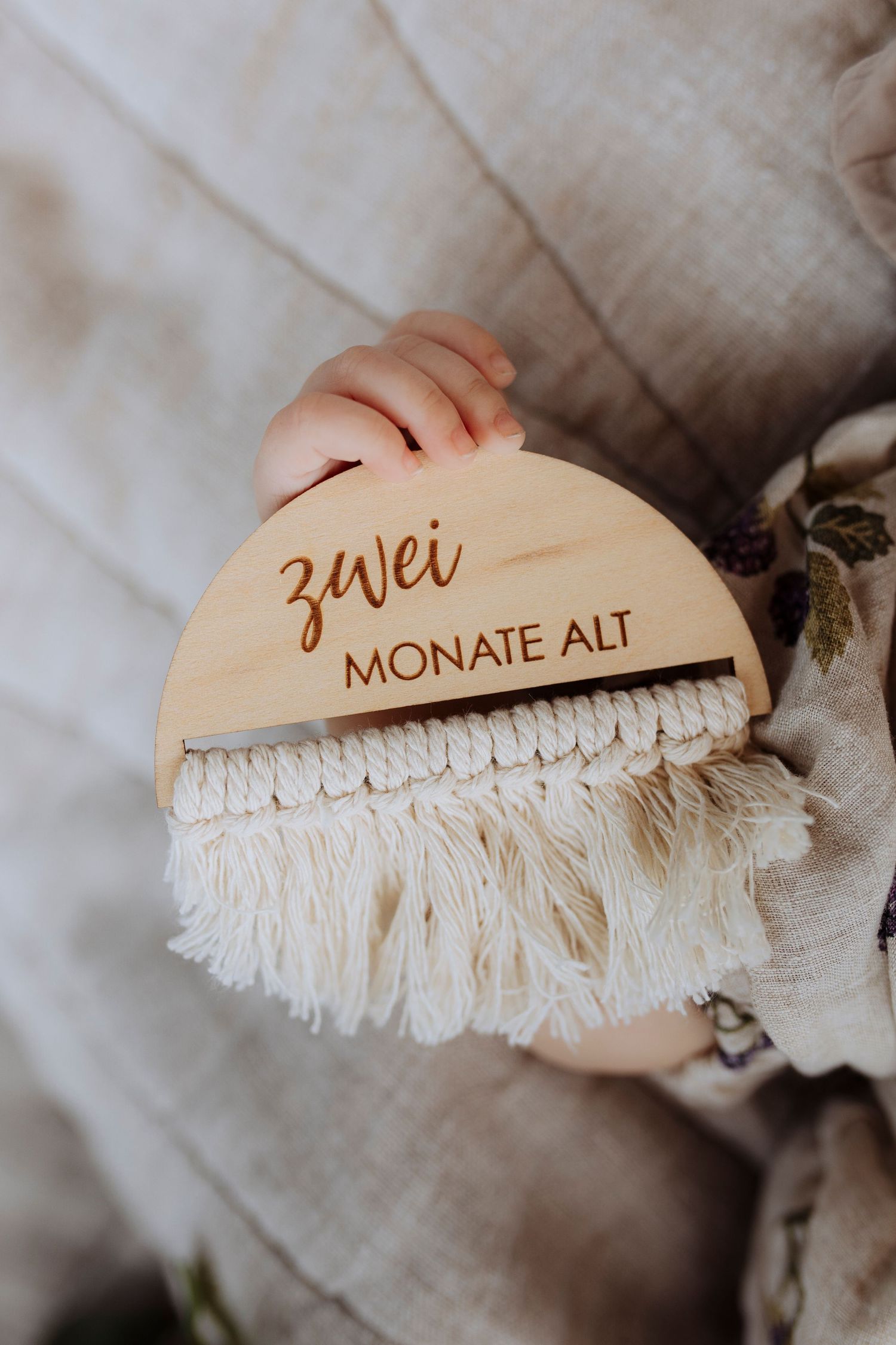 A baby holds a wooden milestone sign reading zwei Monate Alt meaning two months old.