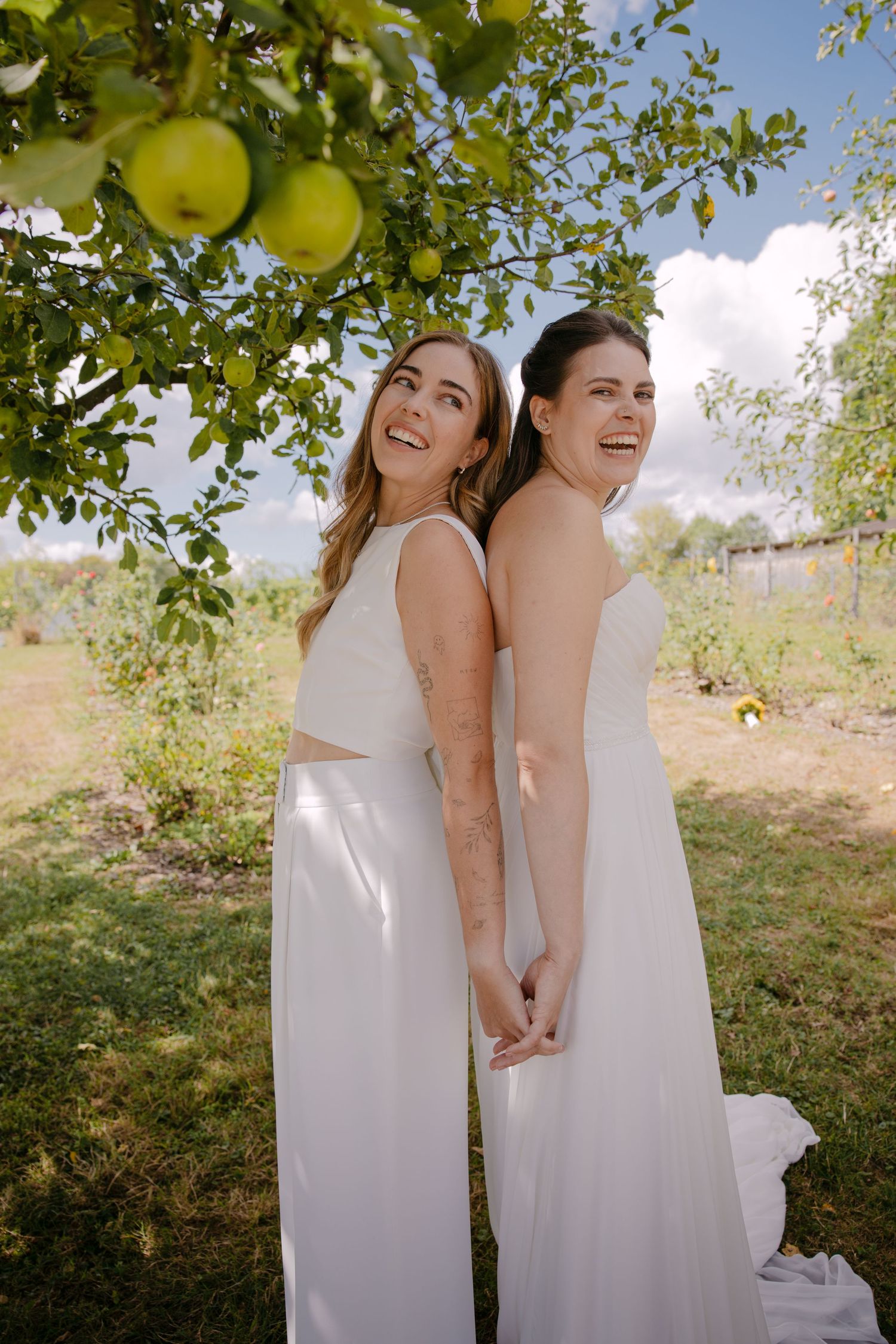 Two brides in white dresses laugh together under an apple tree in a lush garden on a sunny day.