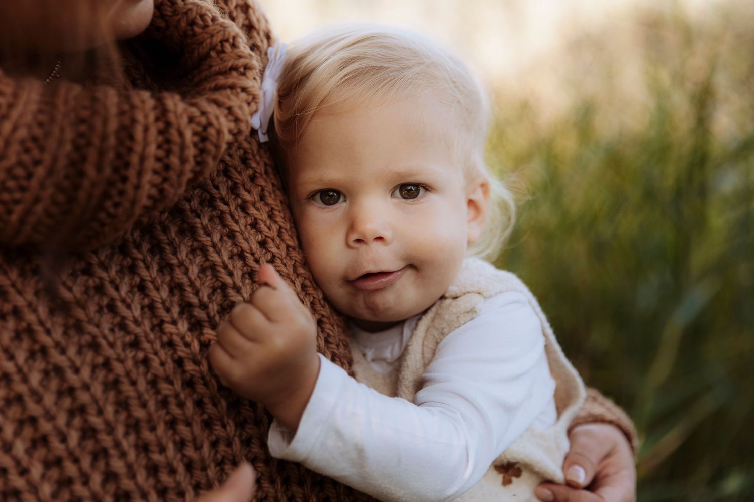 A cute blonde toddler in a white outfit hugs their parent wearing a brown knit sweater outdoors.