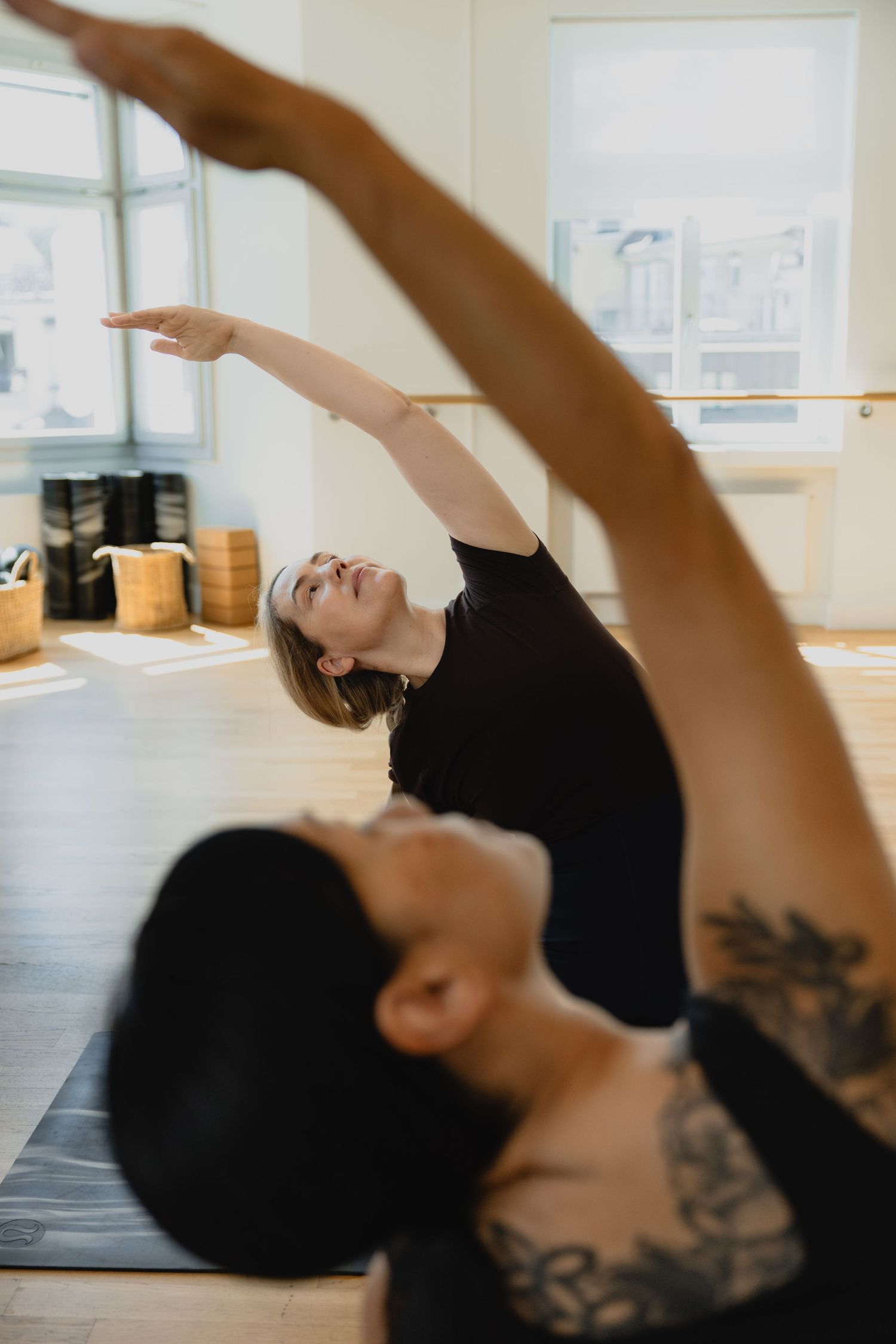 Two women in black practice yoga side stretches in a bright studio with wooden floors and ballet barre.