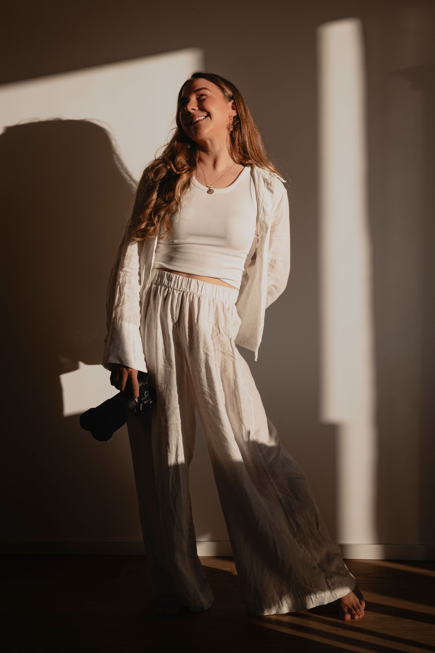Woman in white crop top, linen pants, and blazer smiling in warm sunlight indoors.
