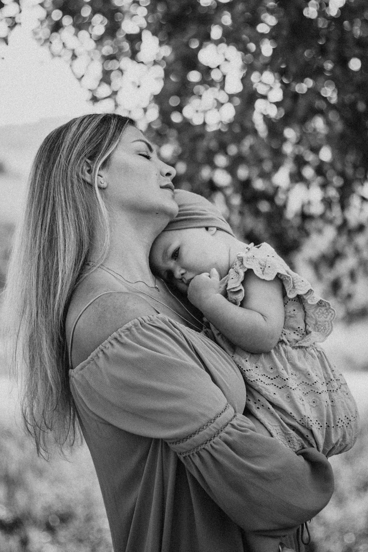 Black and white photo of a mother kissing her sleeping baby outdoors under trees.