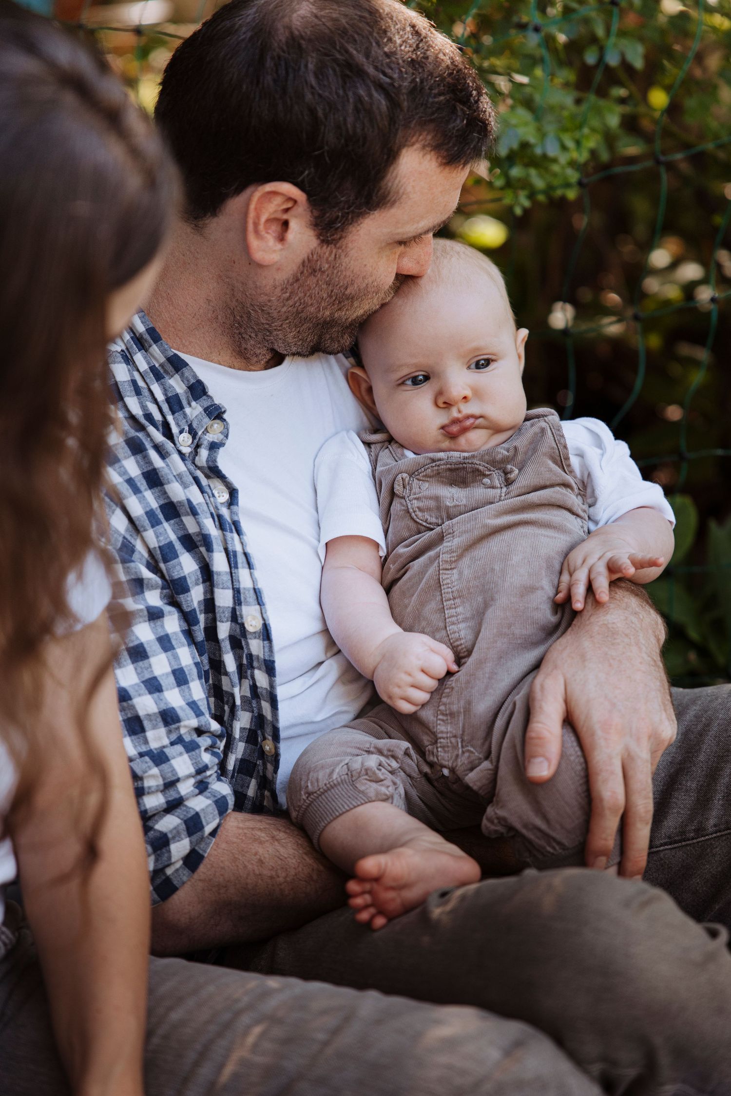 Father kissing his baby's head while holding infant on his lap in a sunny garden setting.